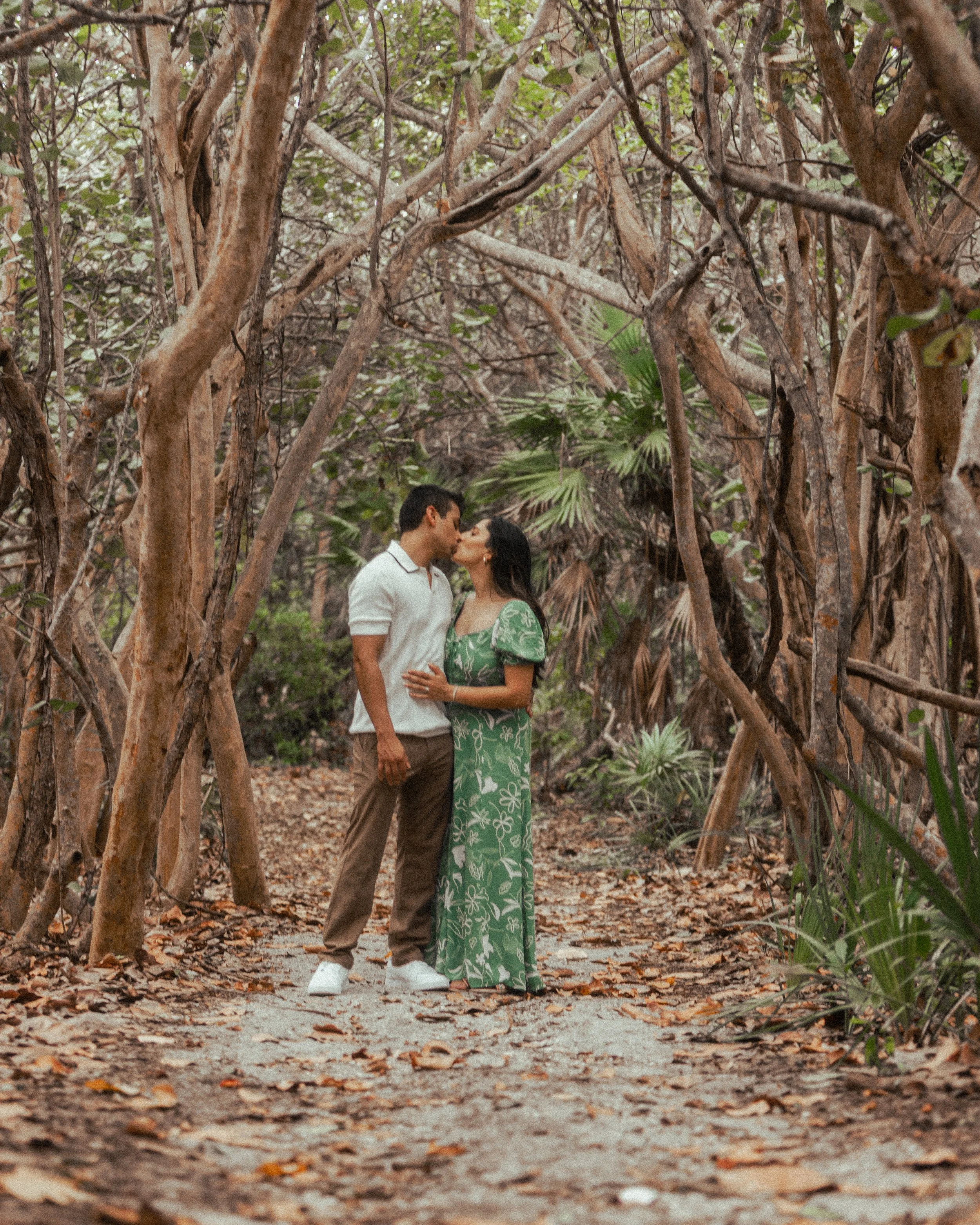 A couple kissing in a wooded area with twisted trees and fallen leaves on the ground.