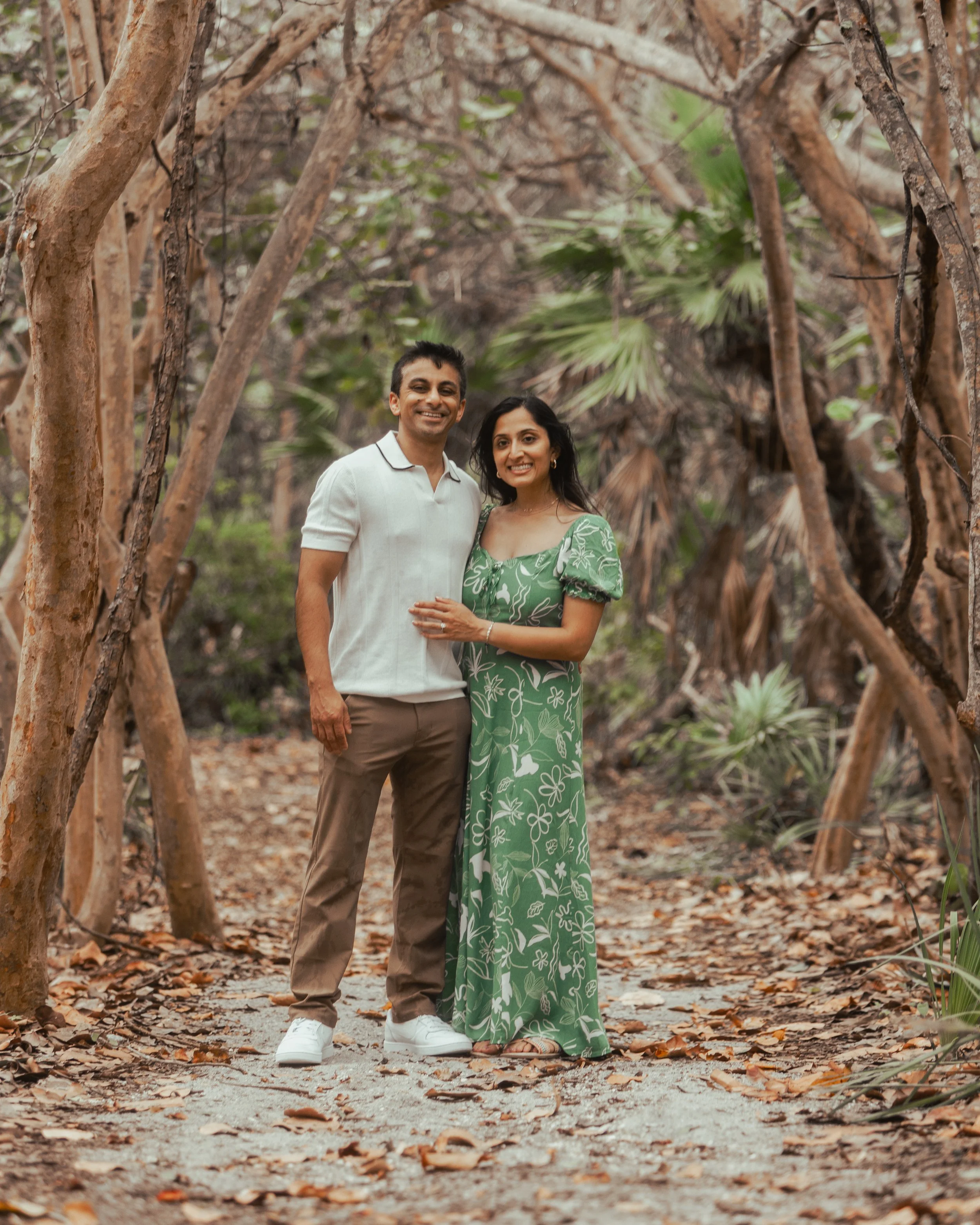A smiling couple standing together in a wooded area with brown and green trees and fallen leaves on the ground.