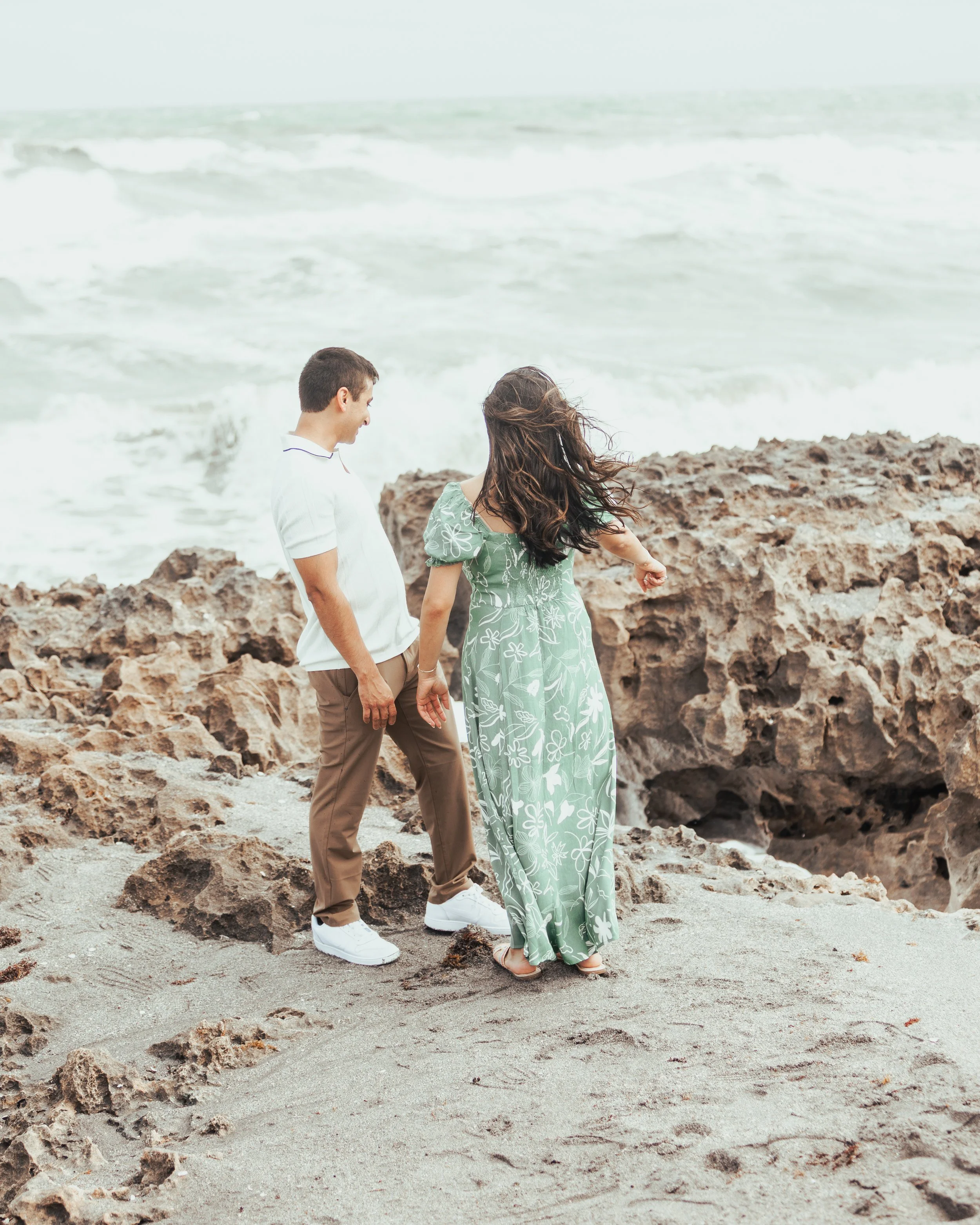 A man and woman standing on a rocky beach near the ocean, holding hands and looking at the water, with waves in the background.