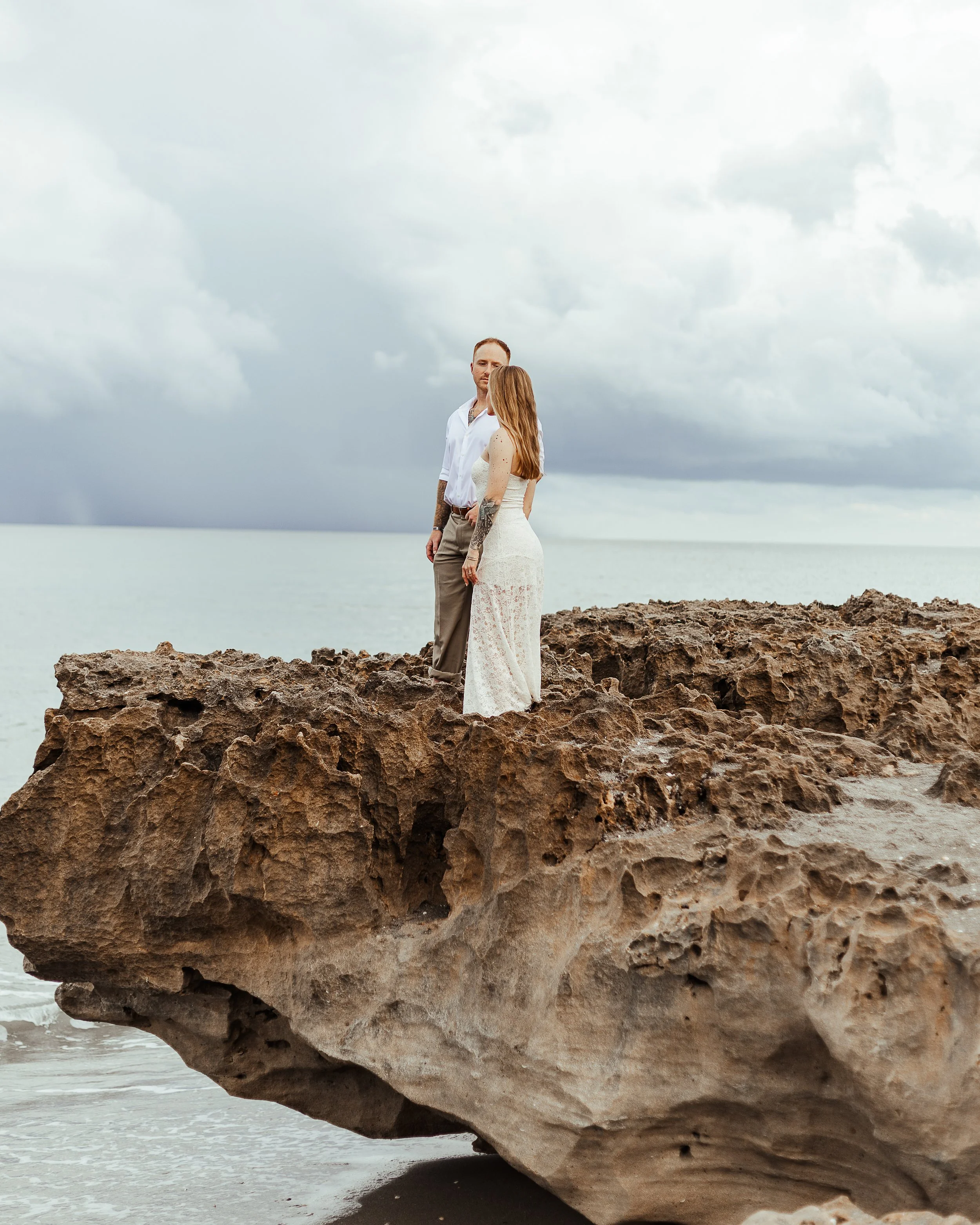 A couple standing on a rocky in Blowing Rocks, FL  facing each other under a cloudy sky, with water visible in the background.