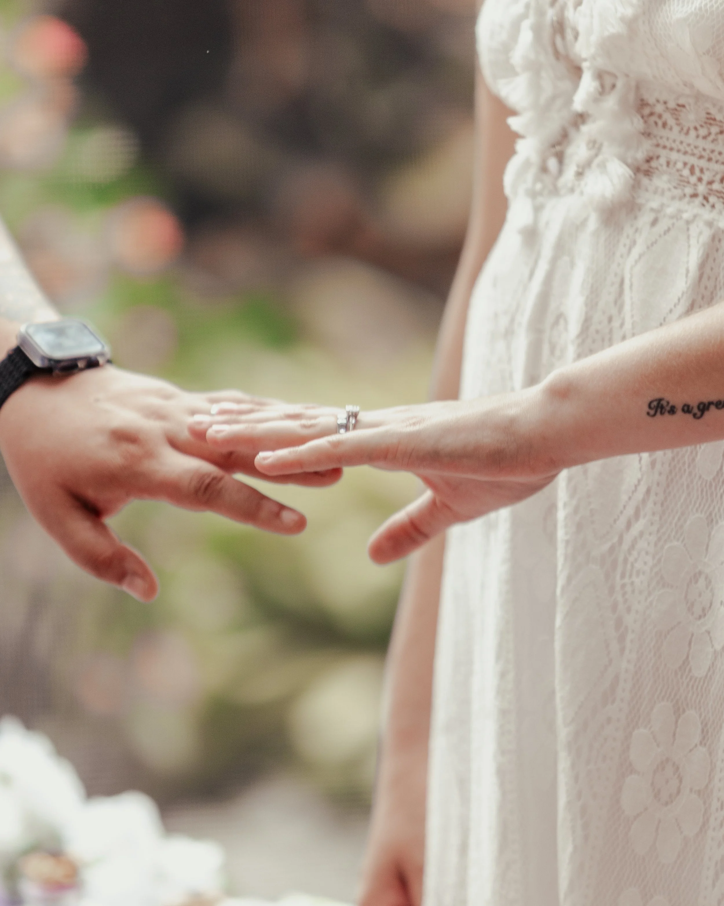 Close-up of a wedding ring exchange between two people, one wearing a white lace dress and a watch, with blurred background.