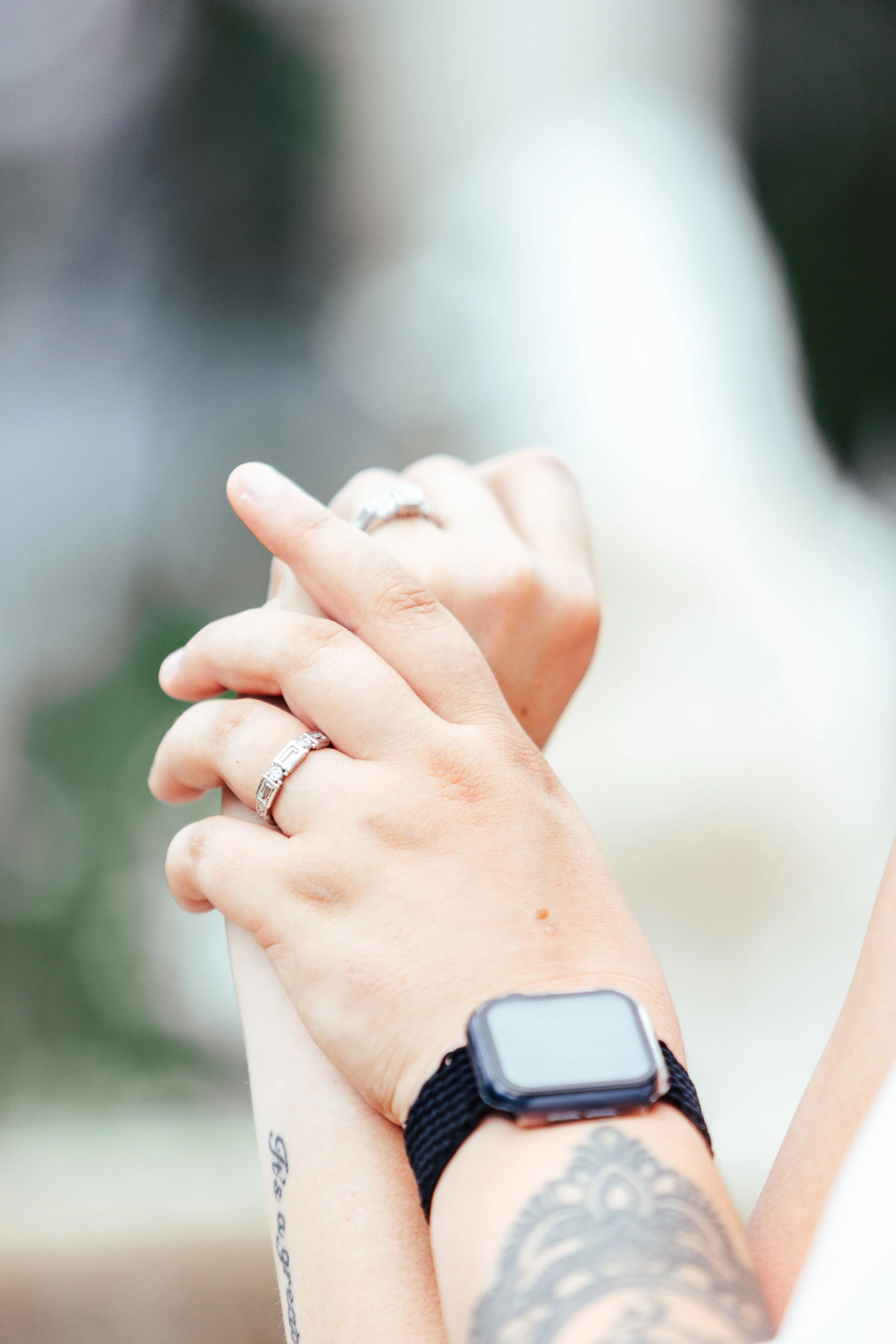 Close-up of two hands clasped together, displaying rings on fingers, with a tattoo on the wrist and a smartwatch on the wrist, against a blurred background.