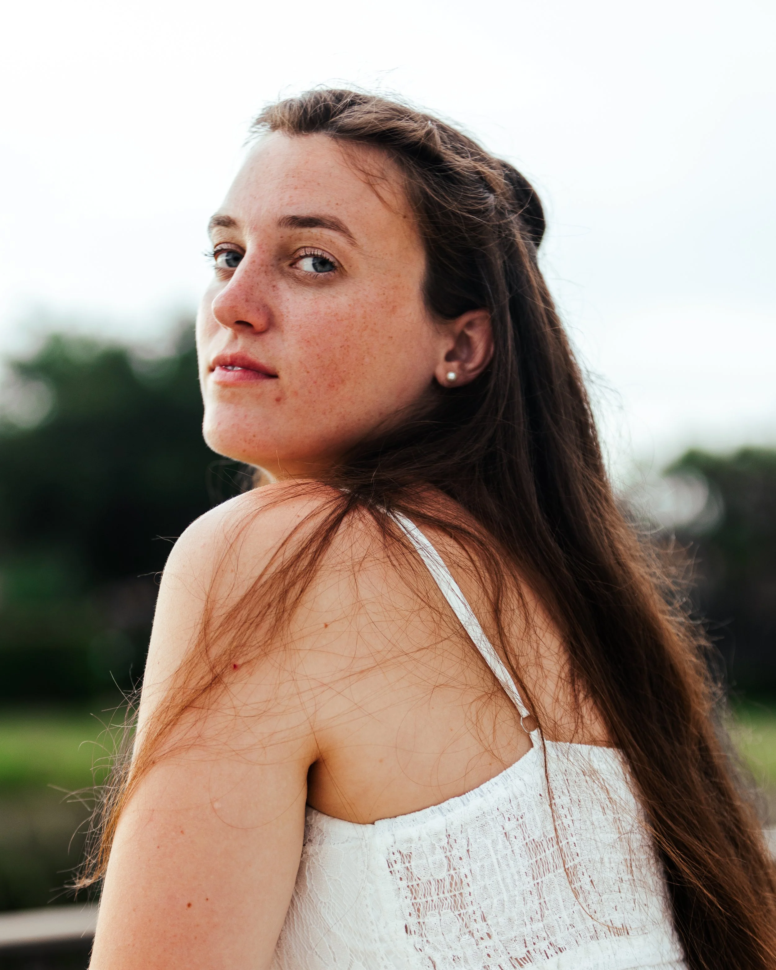A young woman with long brown hair, blue eyes, and freckles, looking over her shoulder outdoors in a white summer dress, with a blurred green landscape in the background.