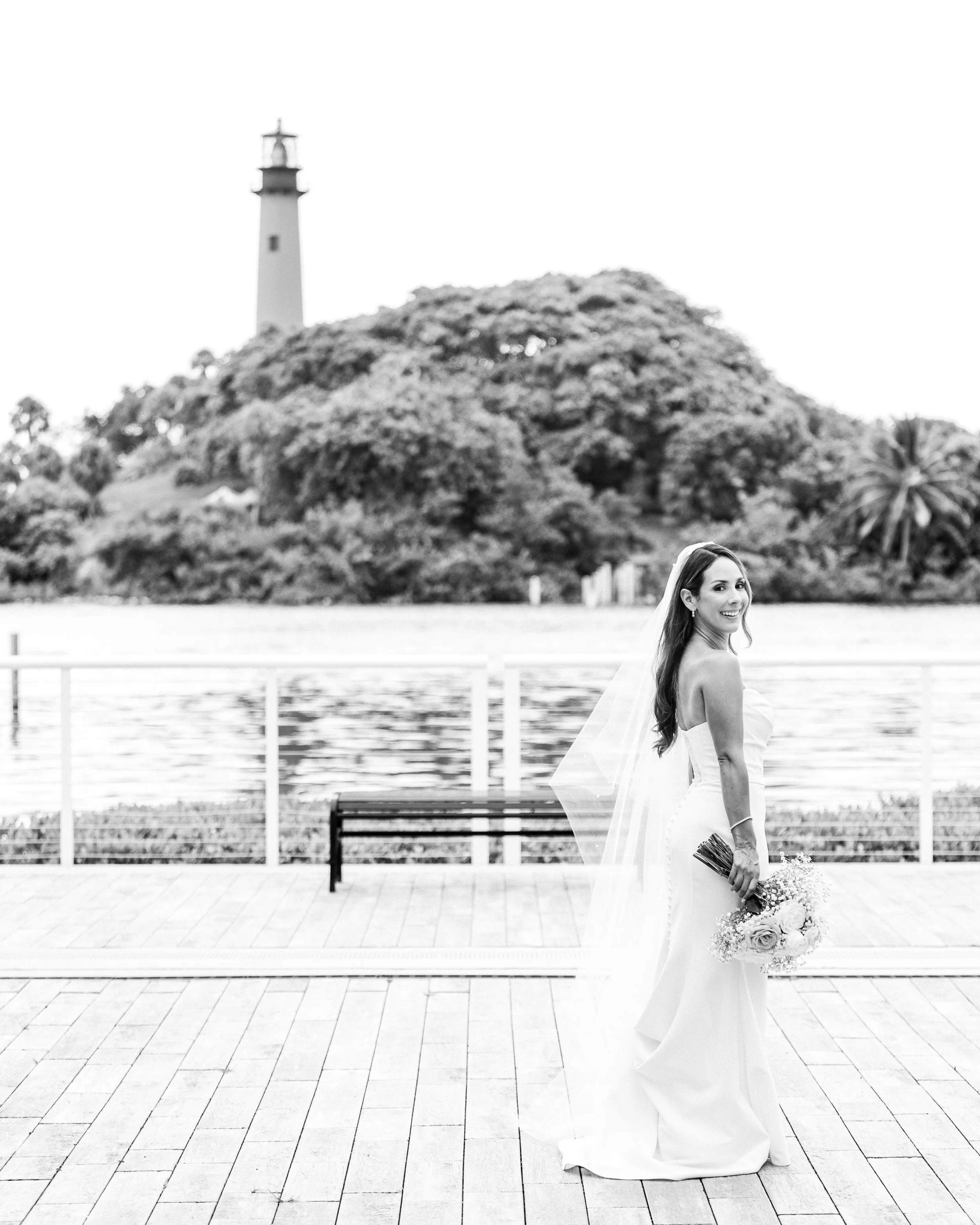A smiling bride in a white wedding dress holding a bouquet in front of a body of water with trees and a lighthouse in the background.