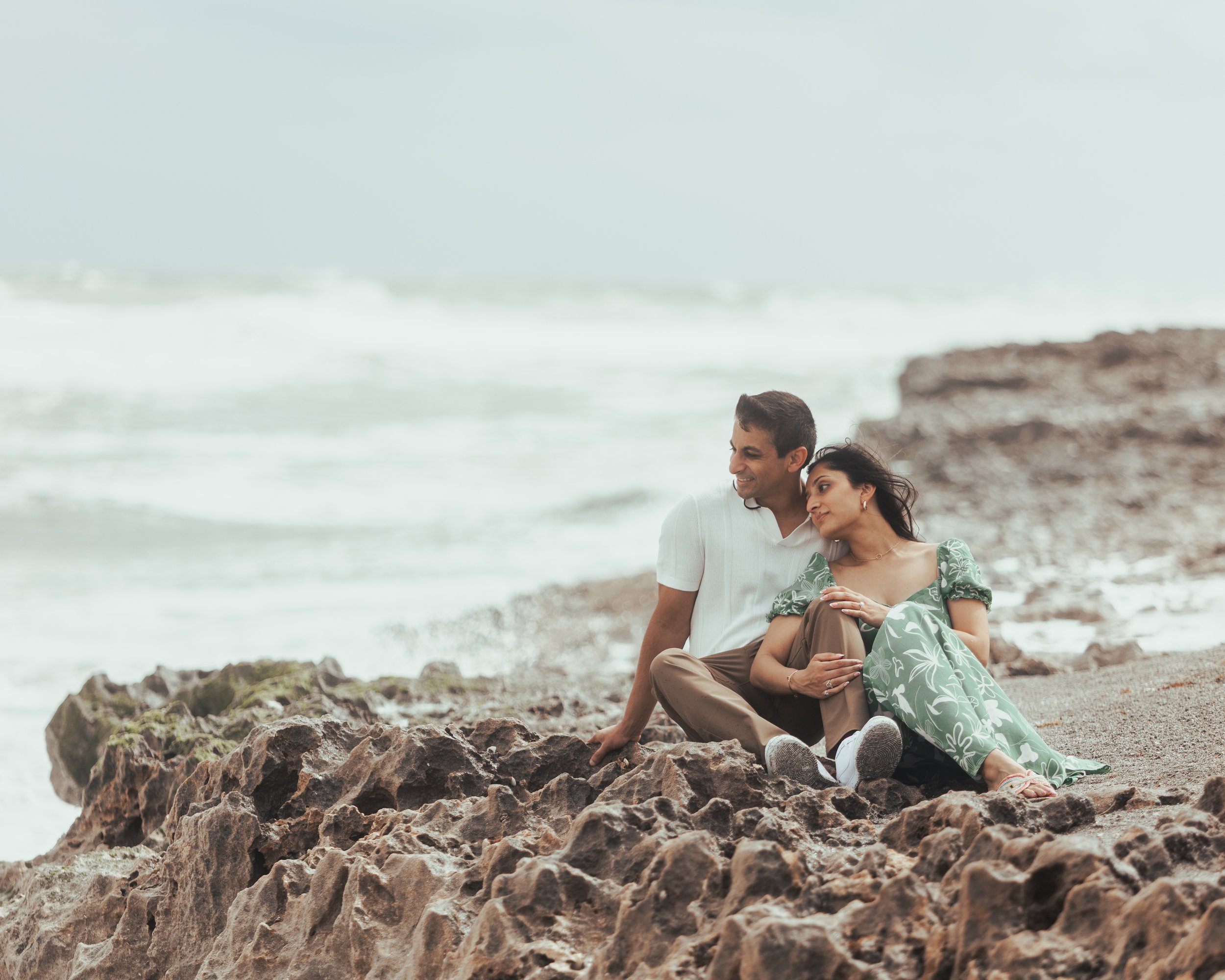 A couple sitting on rocks by the beach, enjoying the ocean view and leaning on each other.
