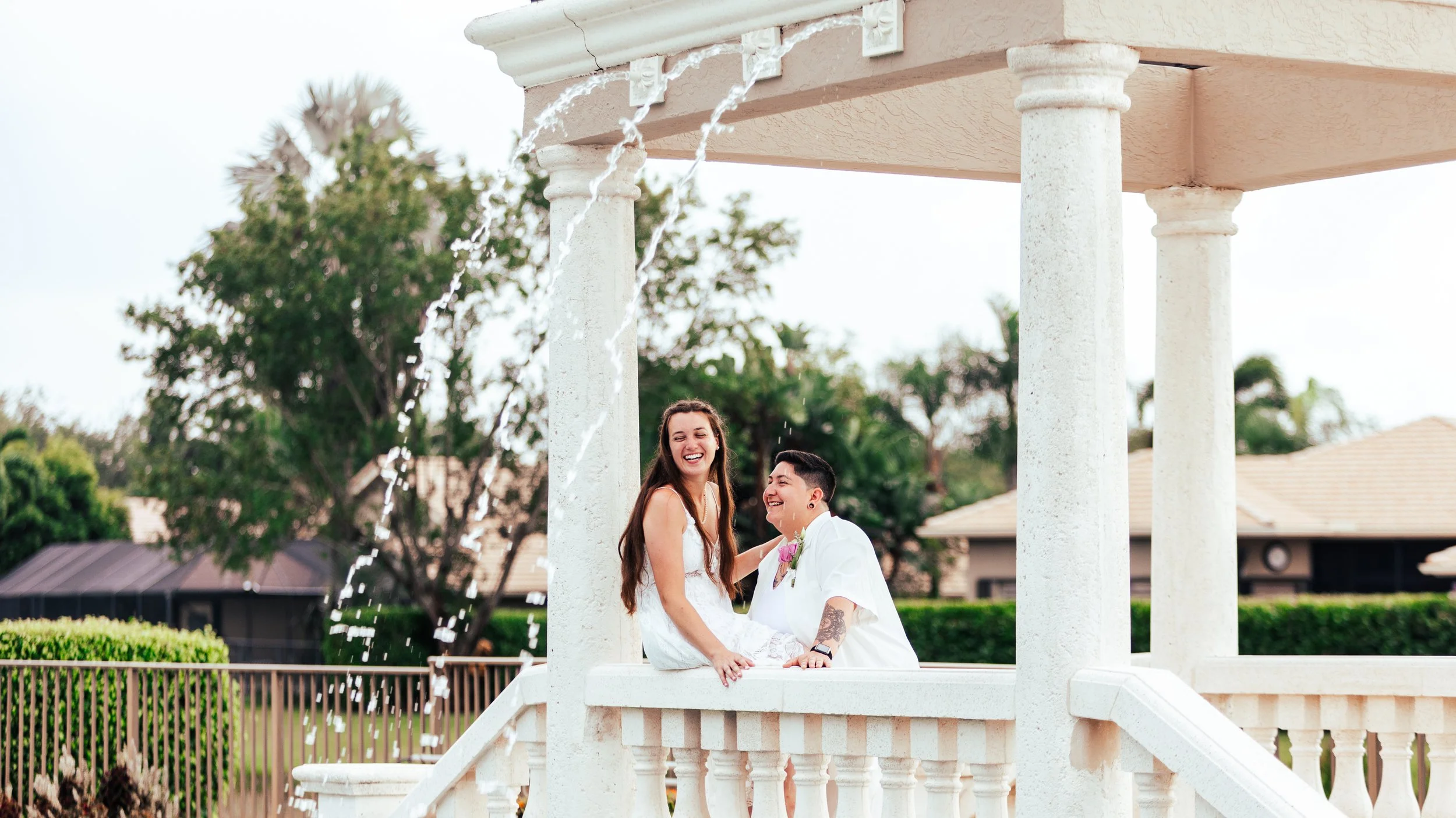 A joyful couple on a wedding day in a gazebo, sharing a moment with water splashing from a fountain, surrounded by greenery and houses in the background.