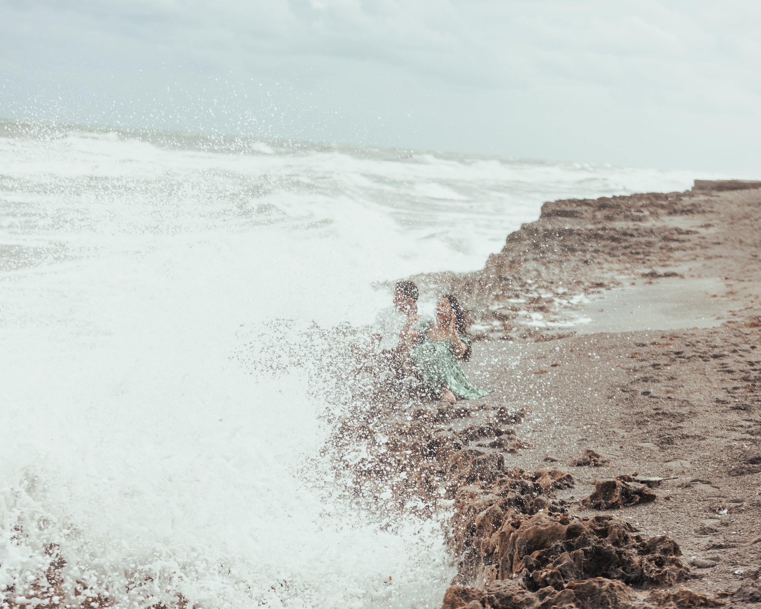 Two children playing near the shoreline as waves crash and splash around them on a cloudy day.