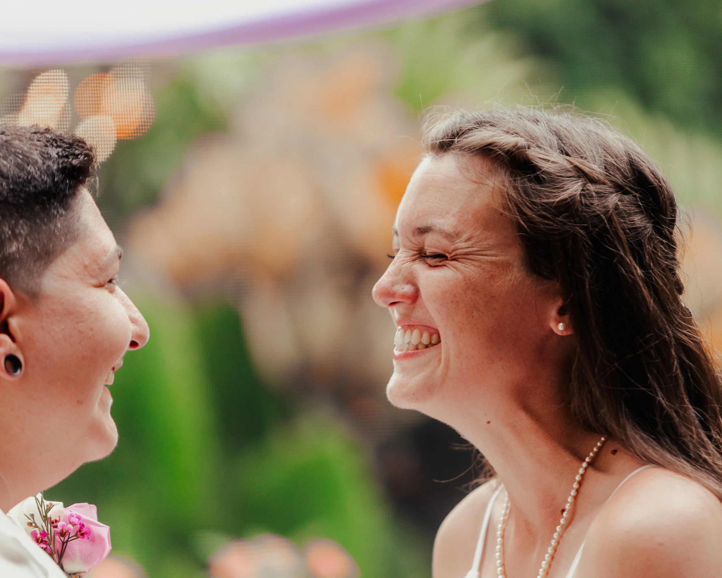Two women facing each other smiling and laughing, one with short curly hair and the other with long wavy hair, outdoors with blurred background of orange and green.