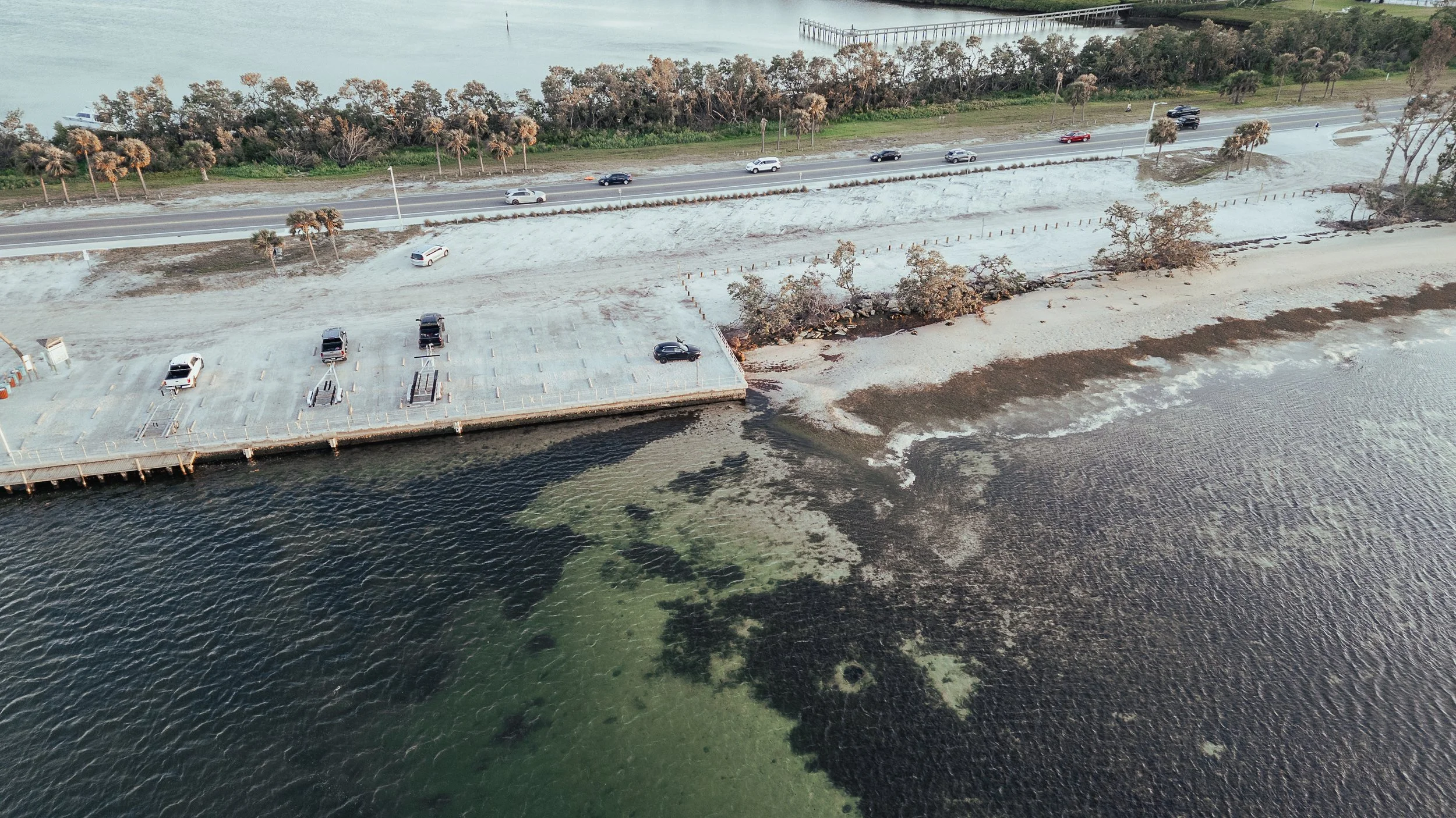 An aerial view of a parking lot next to a body of water, with some cars parked, a few trees, and a road with additional cars in the background.