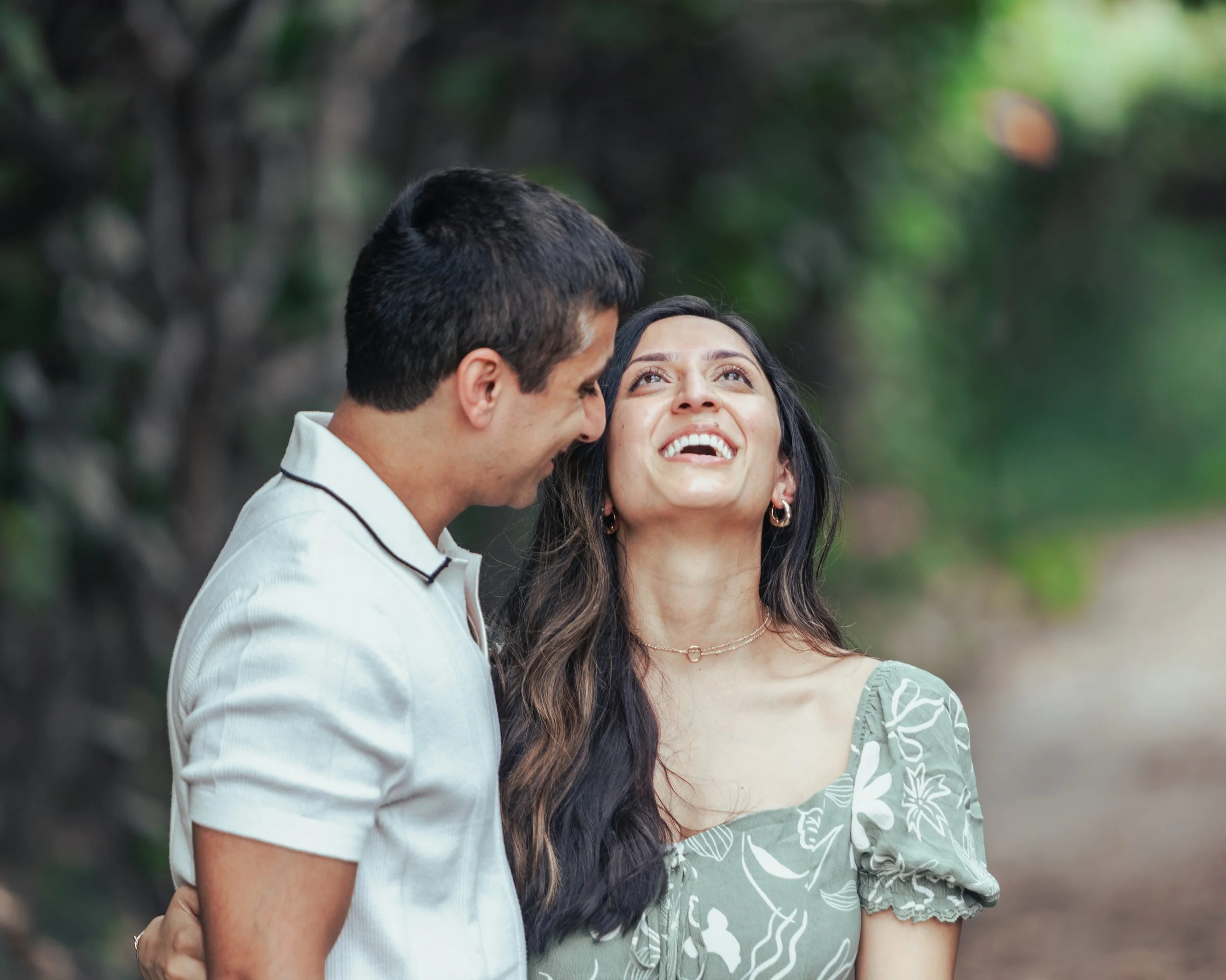 A smiling woman looking up at a man, outdoors with blurred greenery in the background.