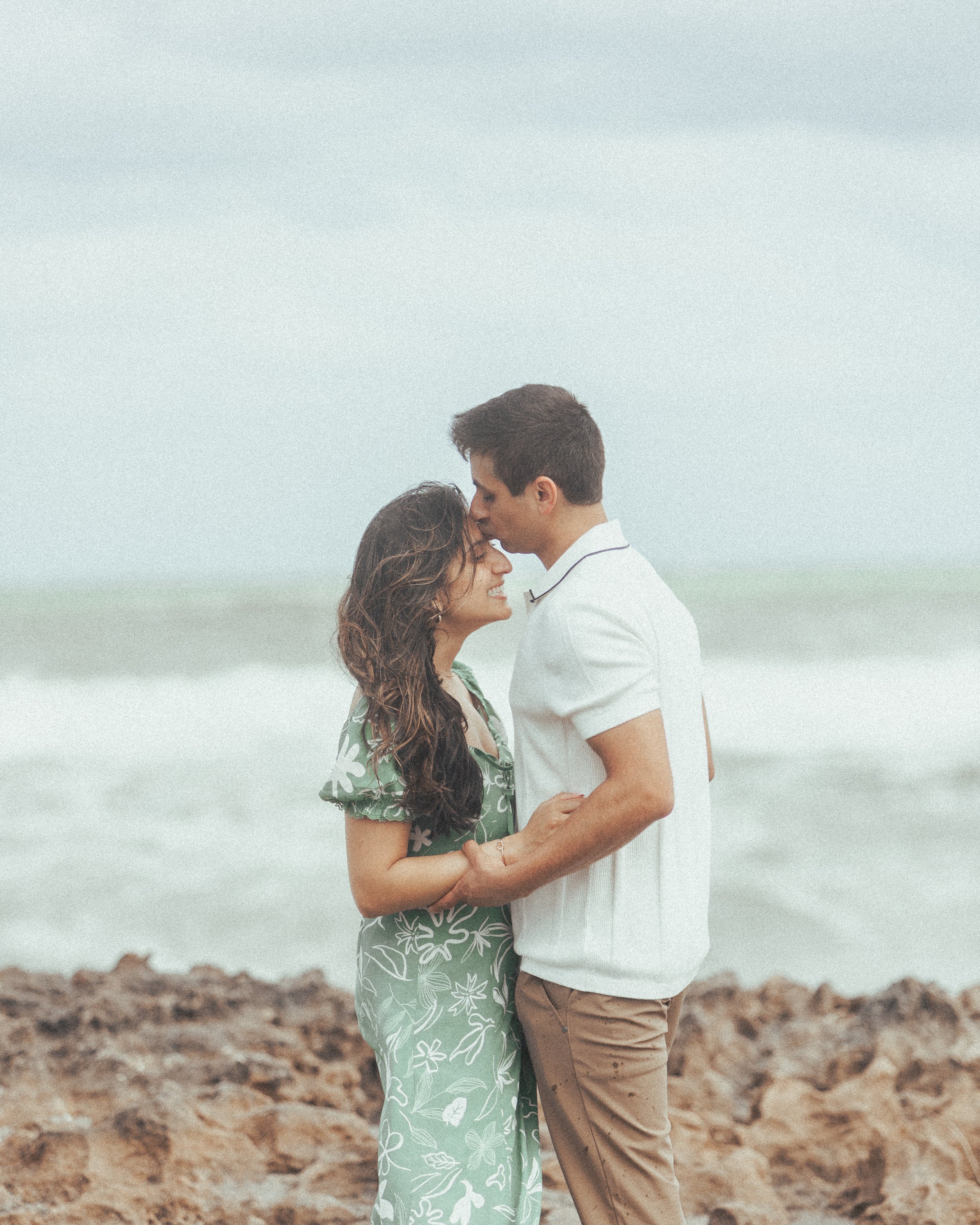 A young couple standing on a rocky beach, facing each other, with the ocean and cloudy sky in the background.