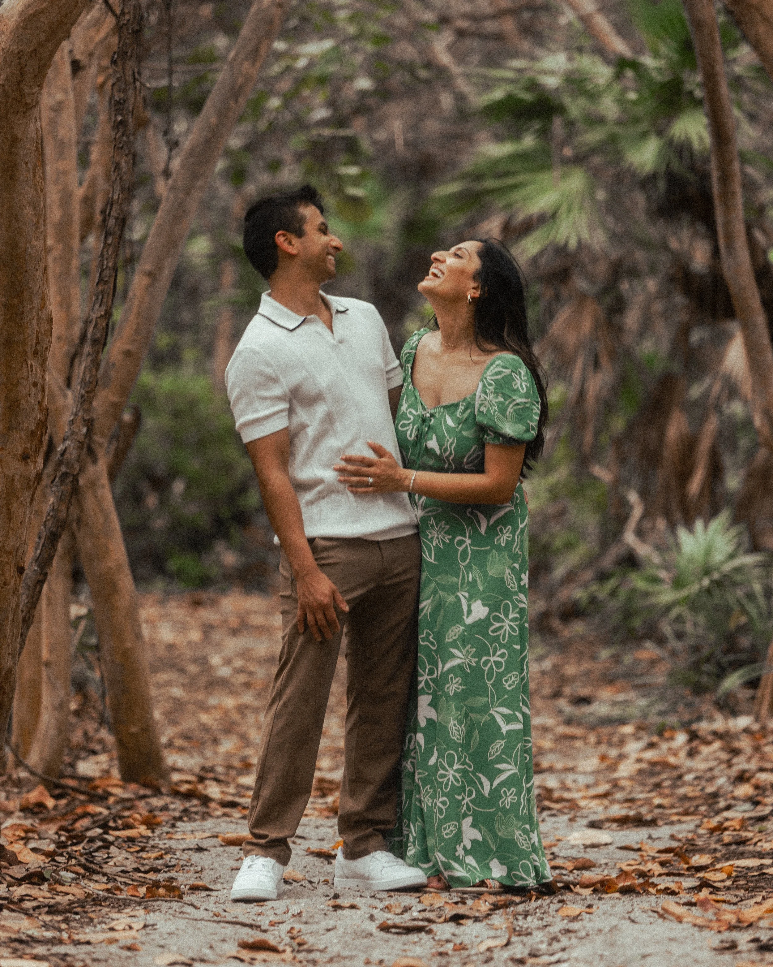 A man and woman standing together in a wooded area, laughing and looking at each other.