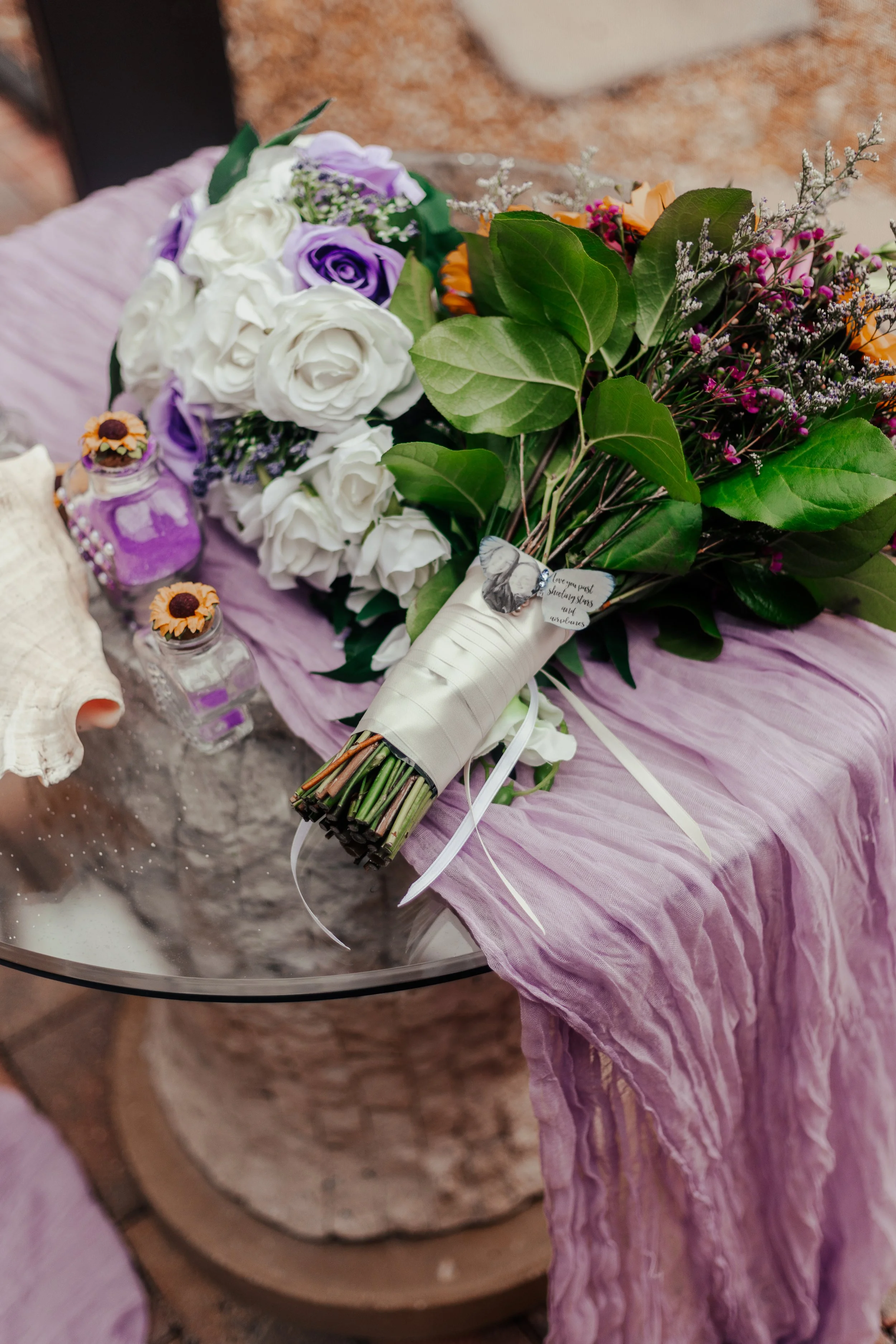 A flower bouquet with white and purple roses, green leaves, and other small flowers lies on a pink fabric draped over a glass table. The bouquet has a tag with a butterfly and a note attached. There are two small jars filled with purple and yellow de