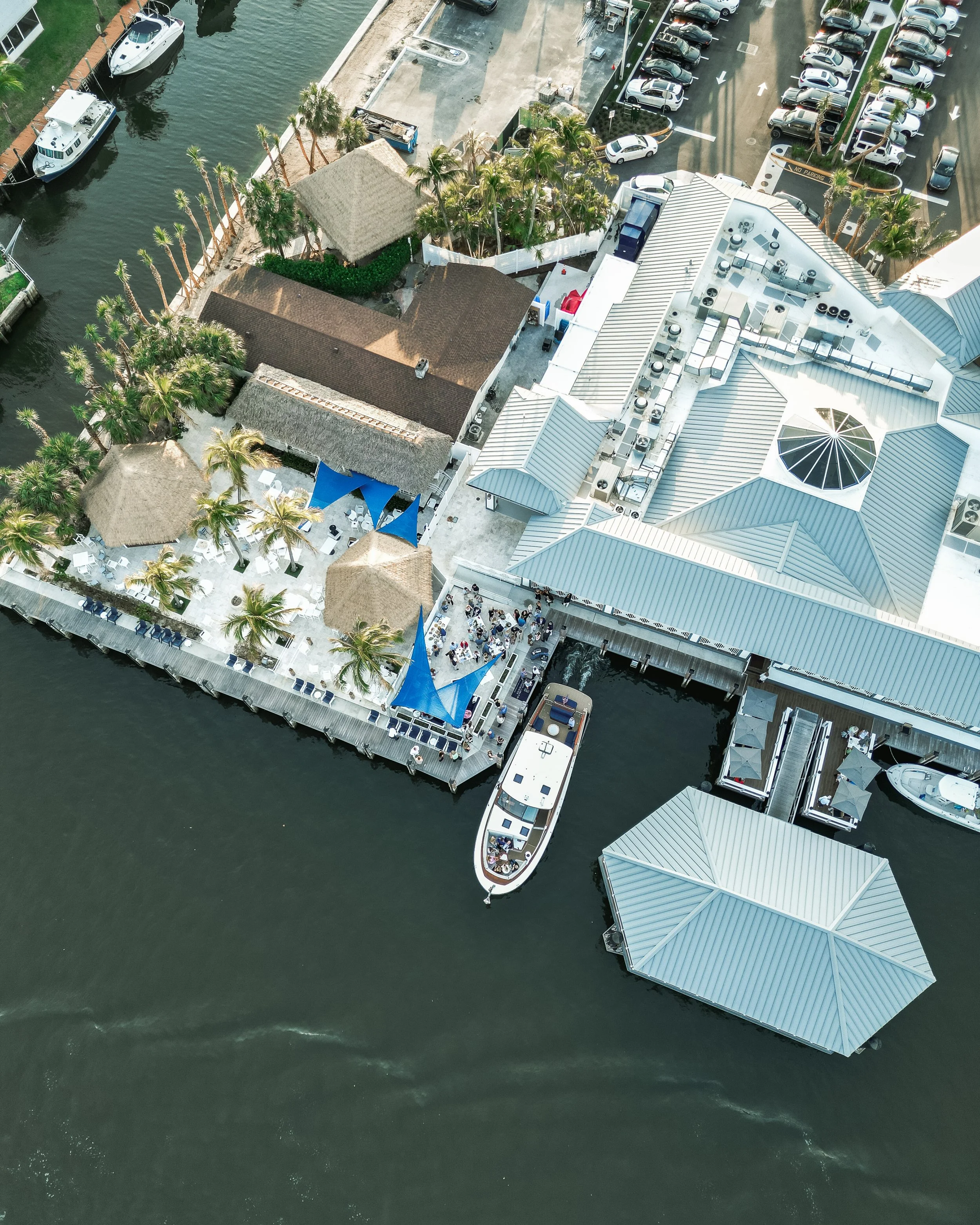 Aerial view of a waterfront area with boats, a deck with blue shade sails, tropical palm trees, a restaurant or event space, parking lot, and buildings with metal roofs.