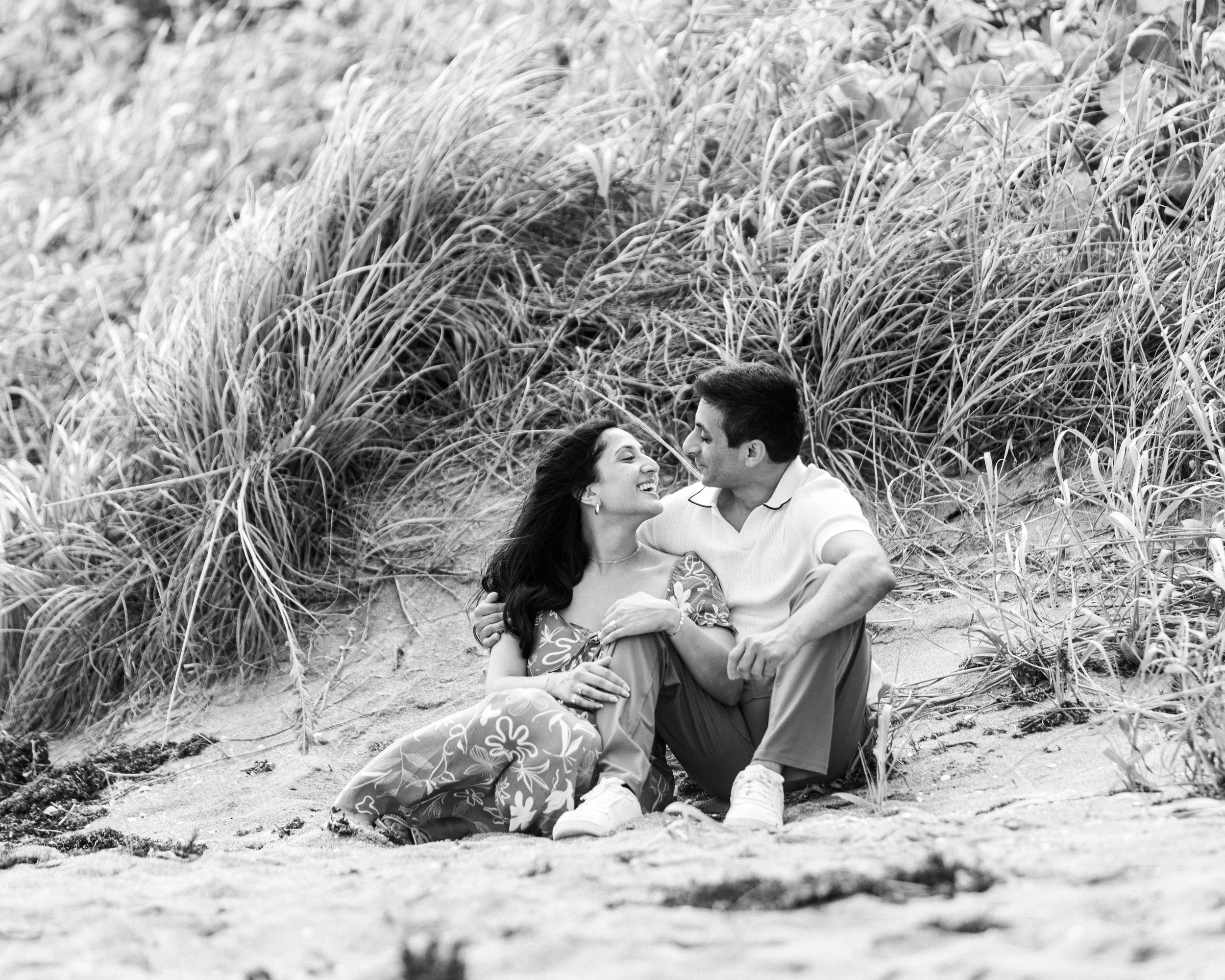 A couple sitting on the sand at the beach, surrounded by tall grass, smiling and looking at each other.