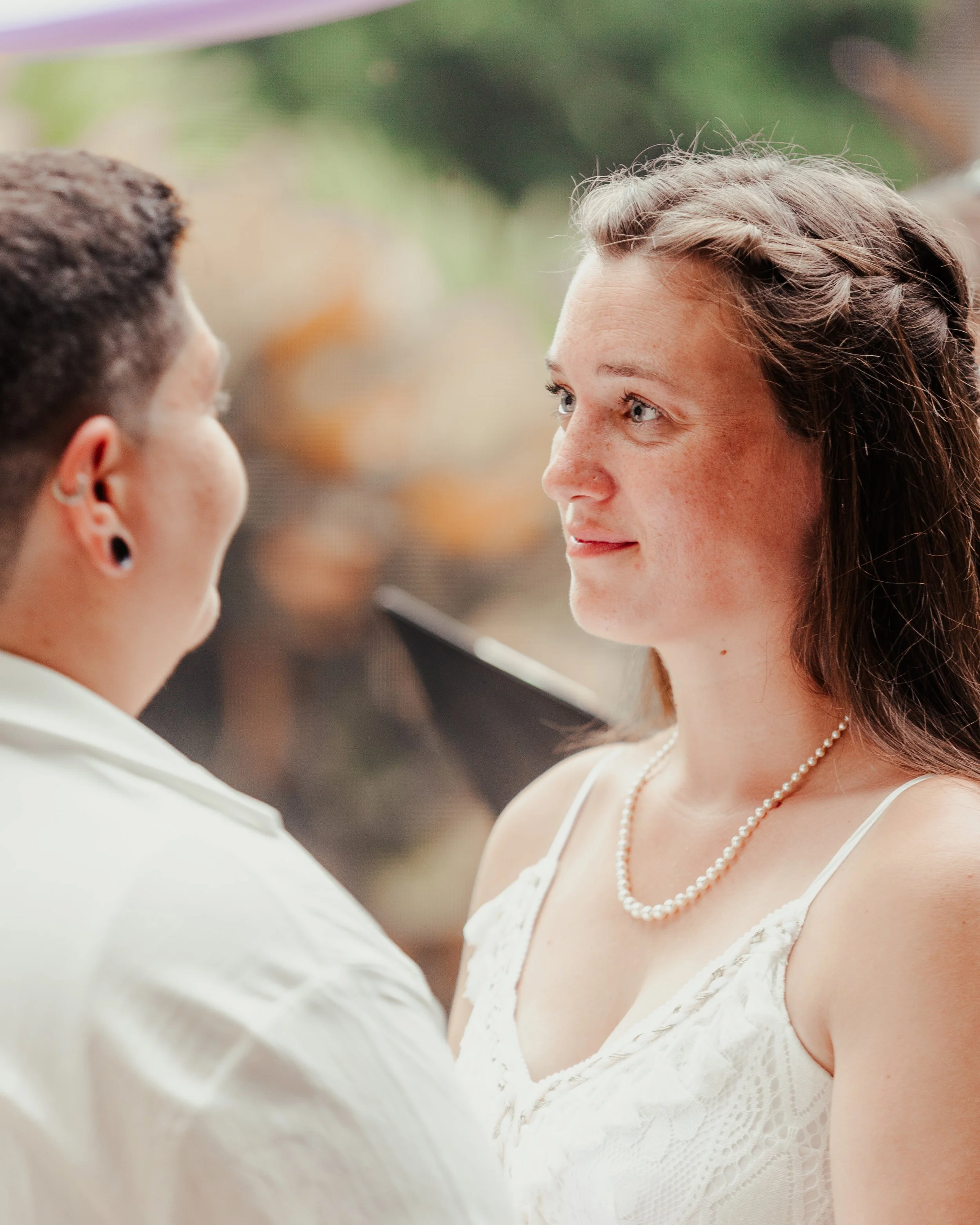 A woman with brown hair wearing a pearl necklace and white dress looks at a person with short brown hair and earrings during a wedding ceremony or similar event.