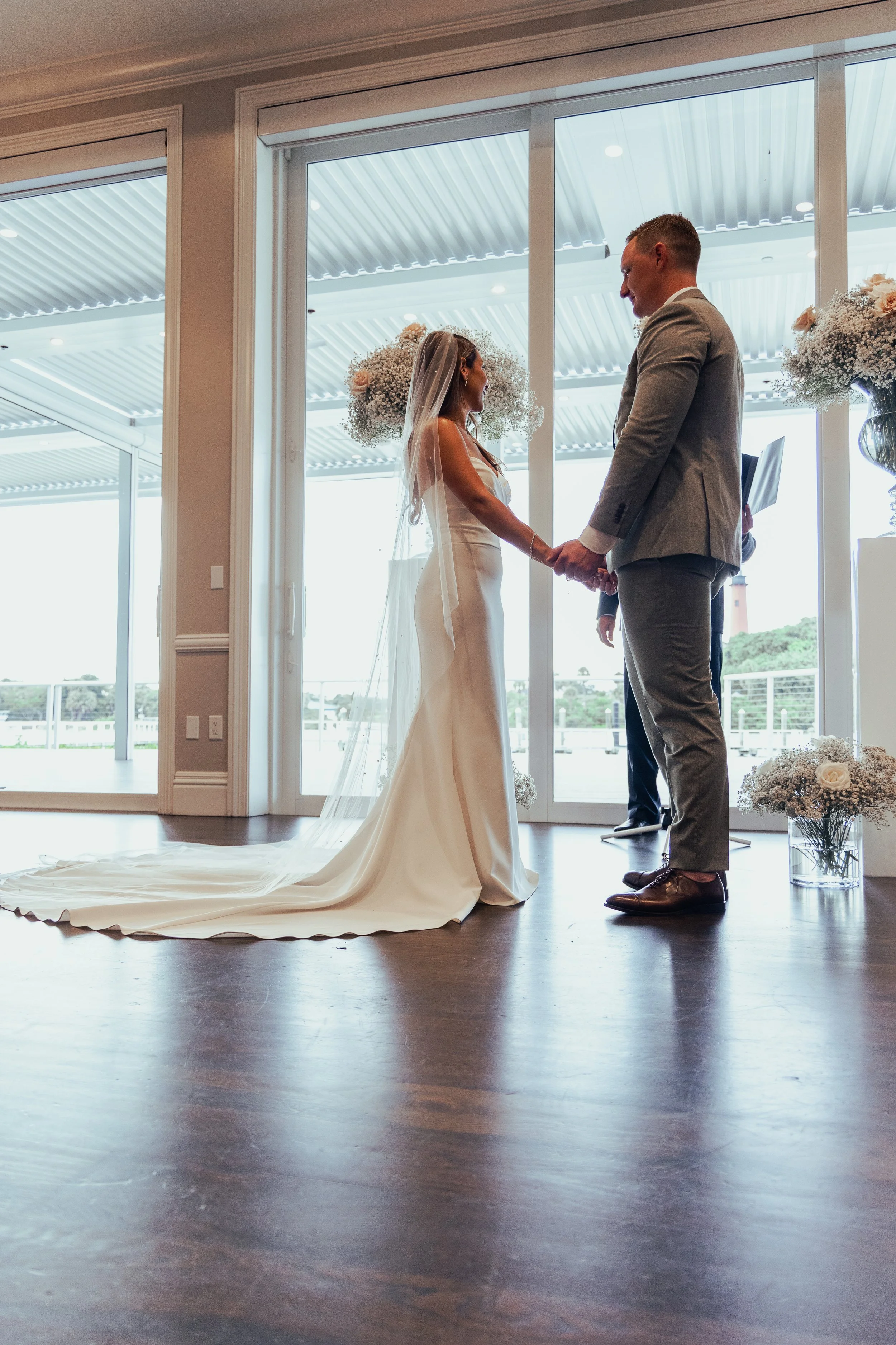A bride and groom holding hands during their wedding ceremony indoors, with large glass doors behind them revealing an outdoor view with trees and a cloudy sky.