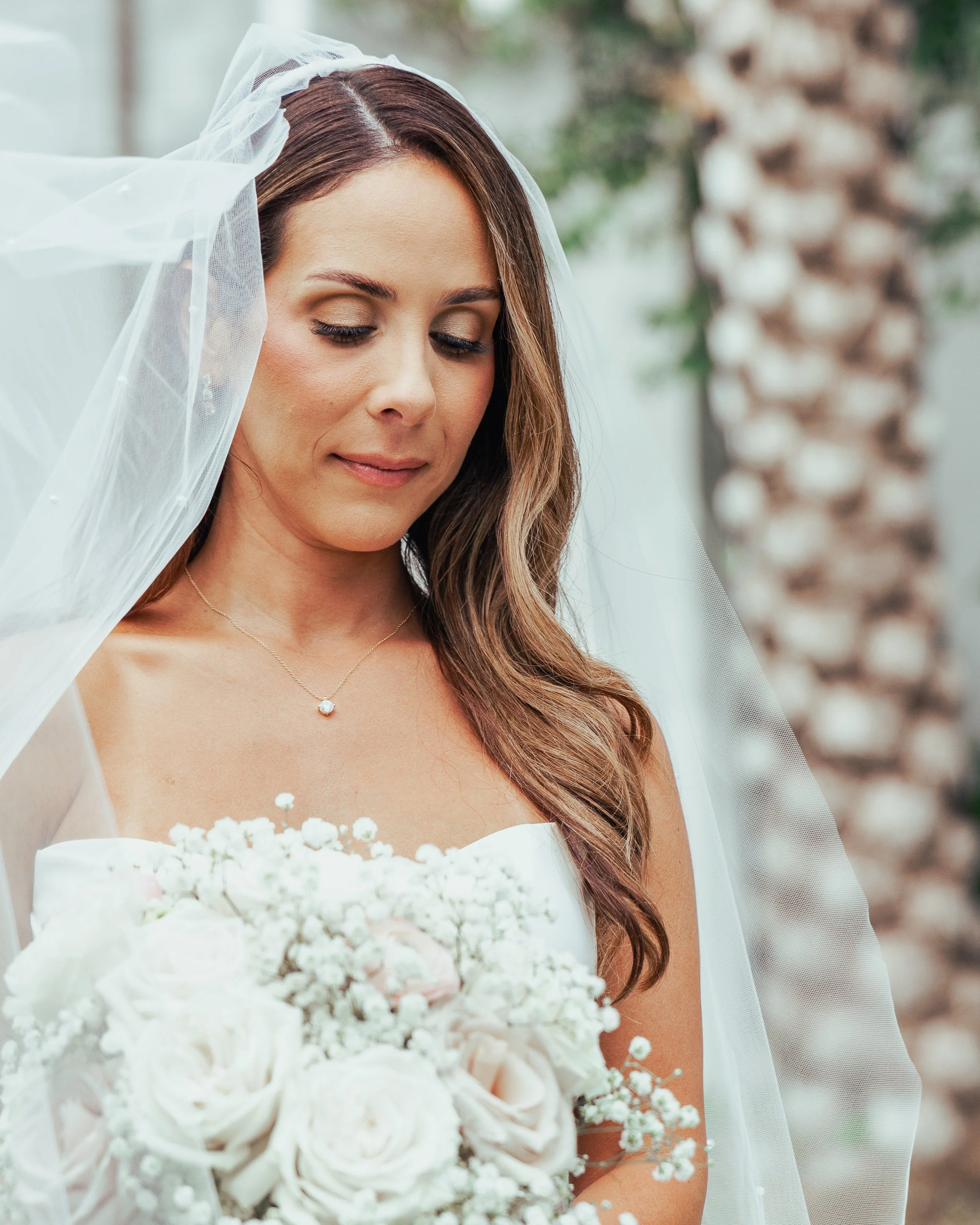 A bride with long brown hair wearing a white strapless wedding dress, a veil, and a necklace, holding a bouquet of white roses and baby's breath, standing outdoors in front of a palm tree.
