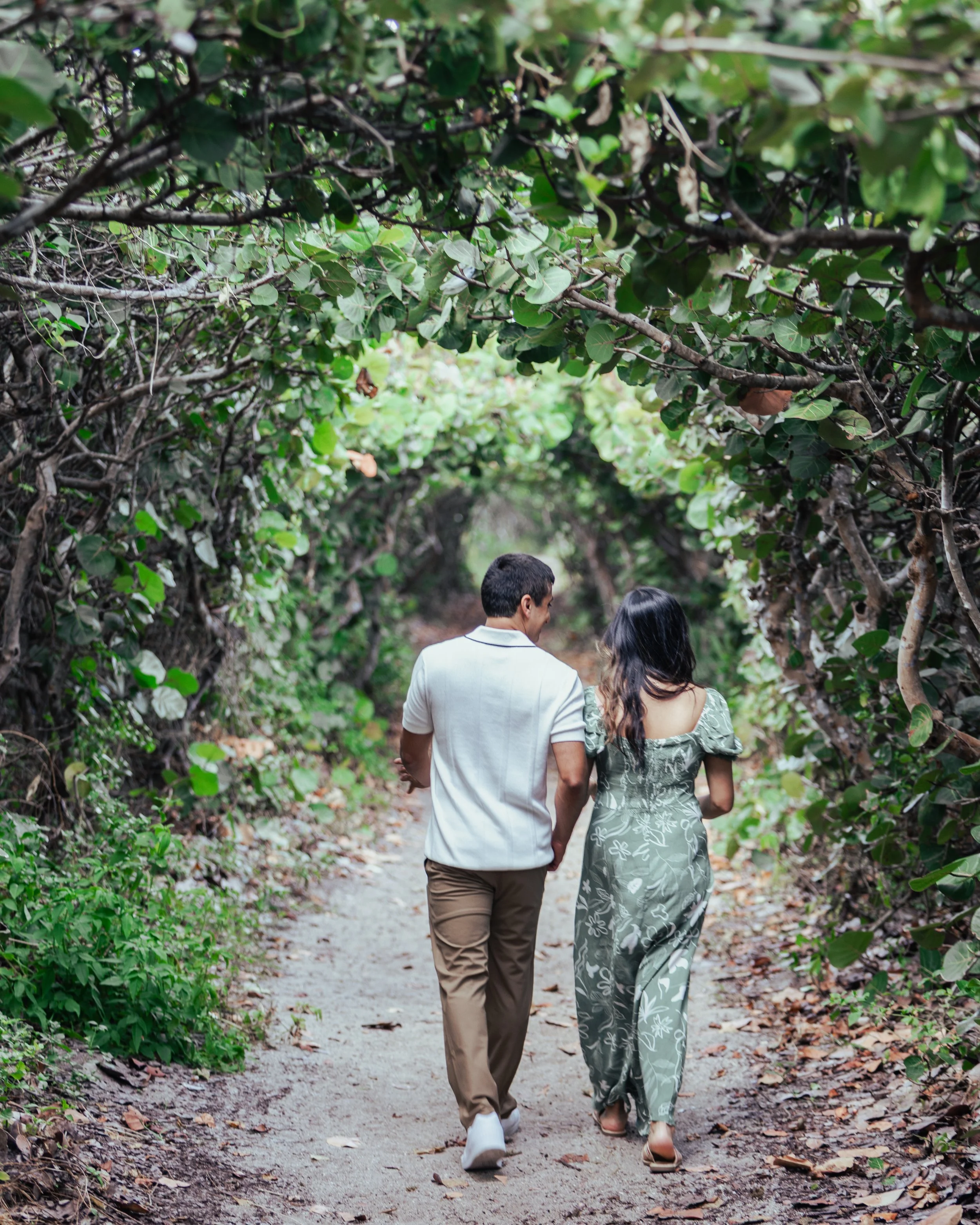 A couple walks hand in hand down a nature trail surrounded by dense greenery and arching tree branches.