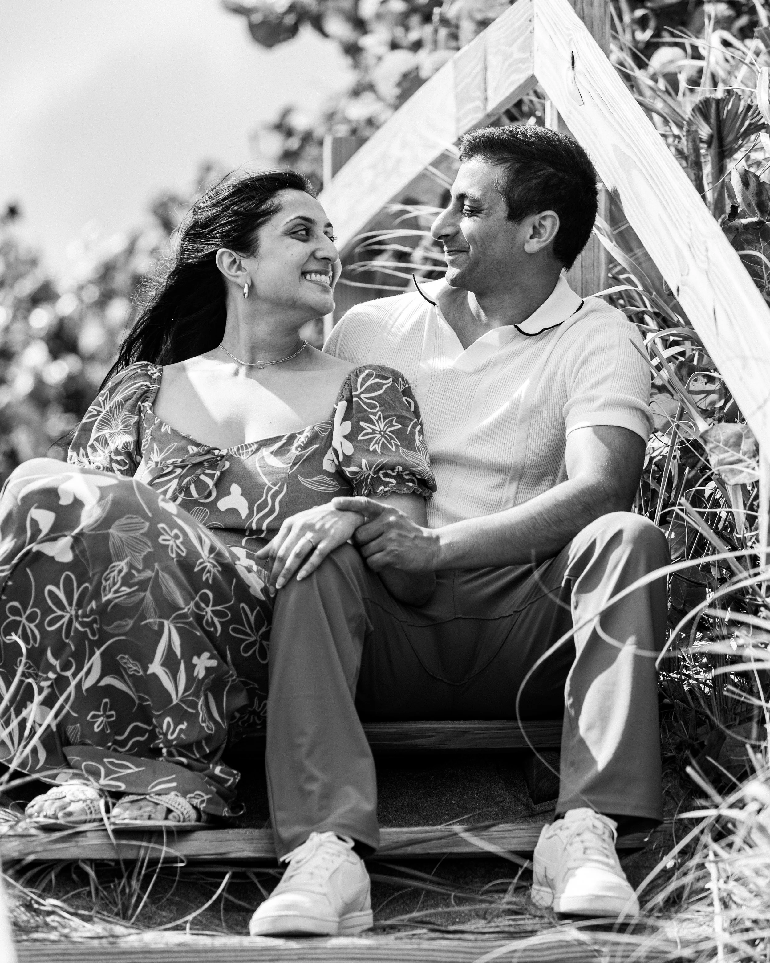 A black and white photo of a couple sitting together outdoors, smiling at each other. The woman wears a floral dress and the man a light-colored polo shirt. They are seated on a bench with plants and wooden beams around them.