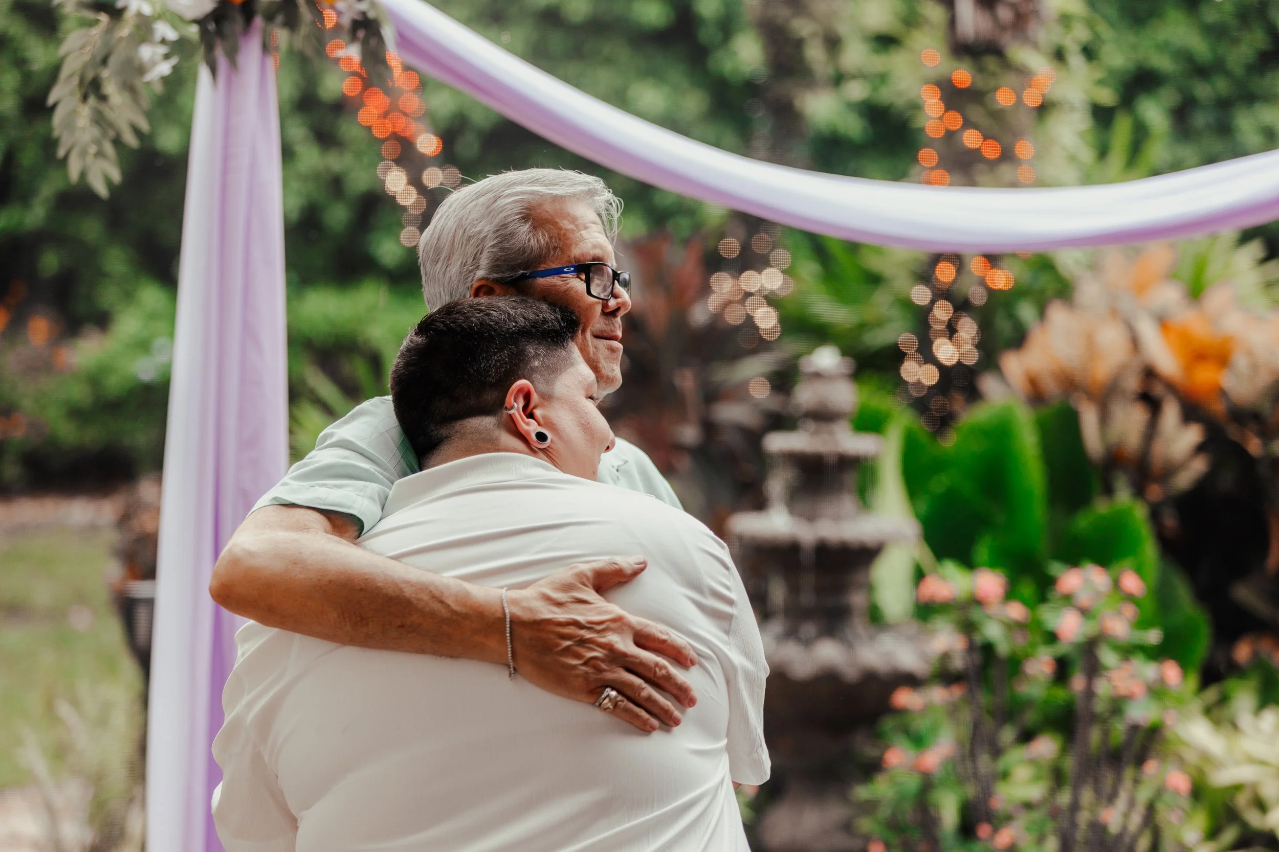 Two people embracing in a hug outdoors at a decorated event, with blurred greenery and string lights in the background.