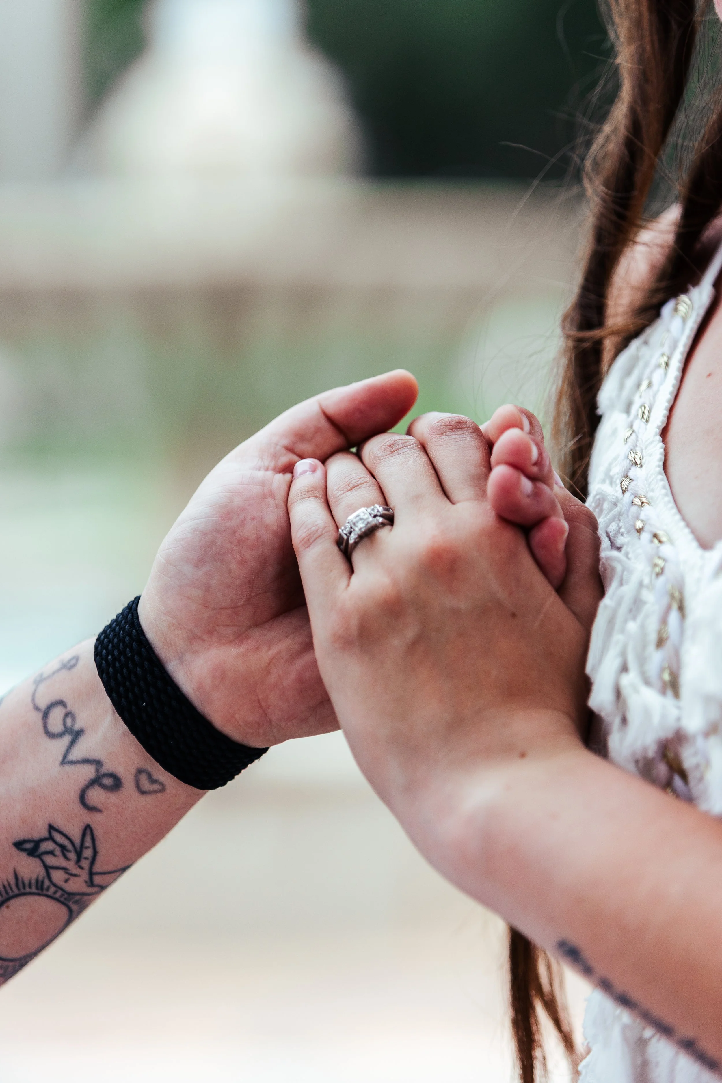Close-up of two people holding hands, one person wearing a wedding ring, tattoos visible on their wrist.