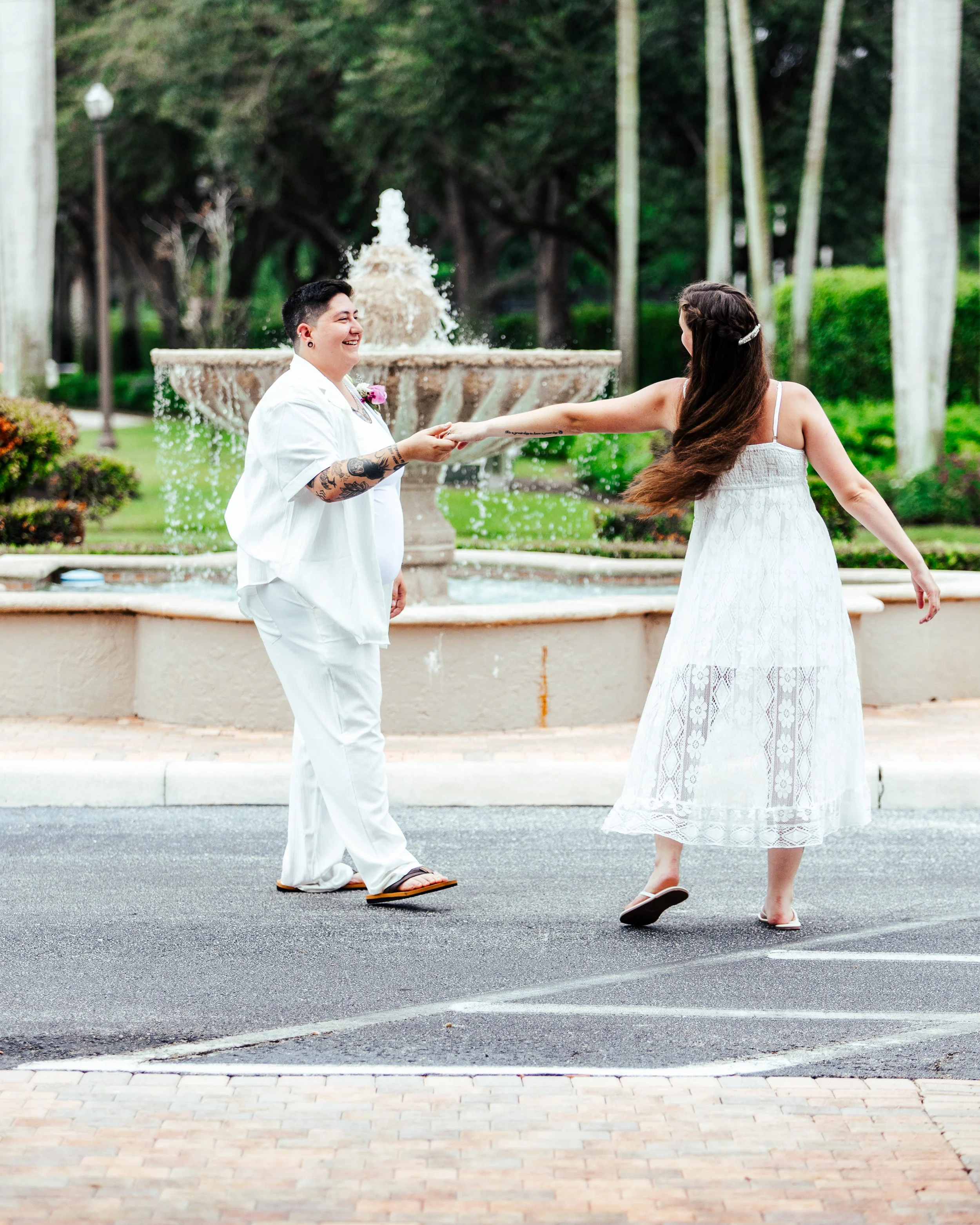 Two women, one with tattoos in white clothing and the other in a white dress, dancing hand in hand in front of a fountain in a park.
