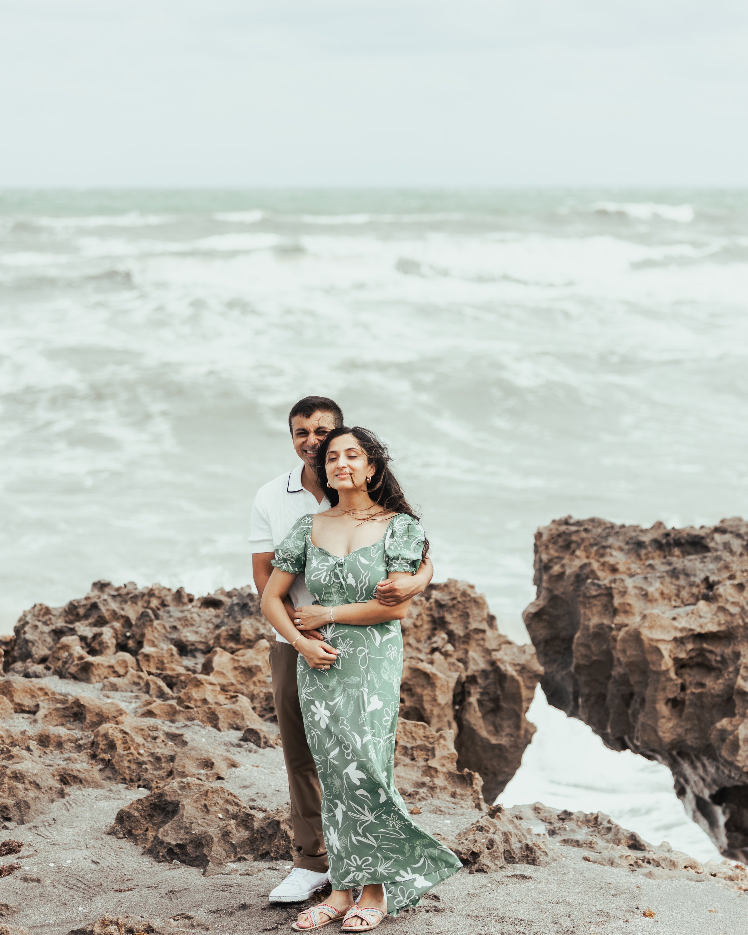A couple stands on rocky beach with ocean waves in the background. The woman wears a green floral dress, and the man wears a white shirt and khaki pants. They embrace, smiling peacefully.