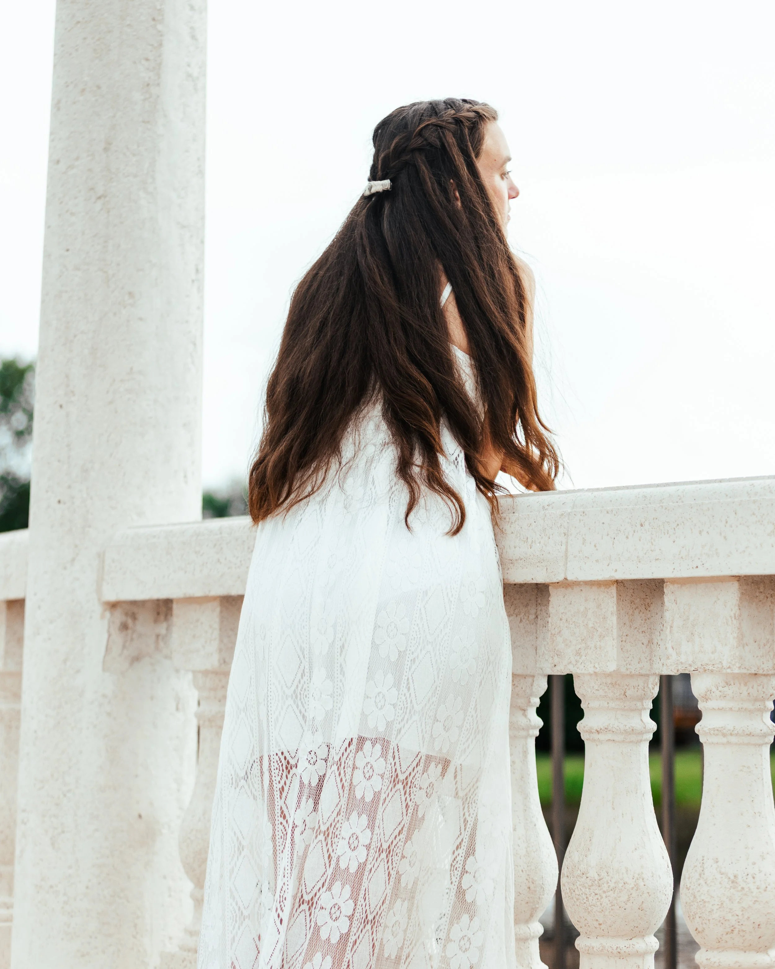 A woman with long brown hair, styled in a braid, leaning on a concrete railing outdoors, wearing a sheer white dress with floral patterns.