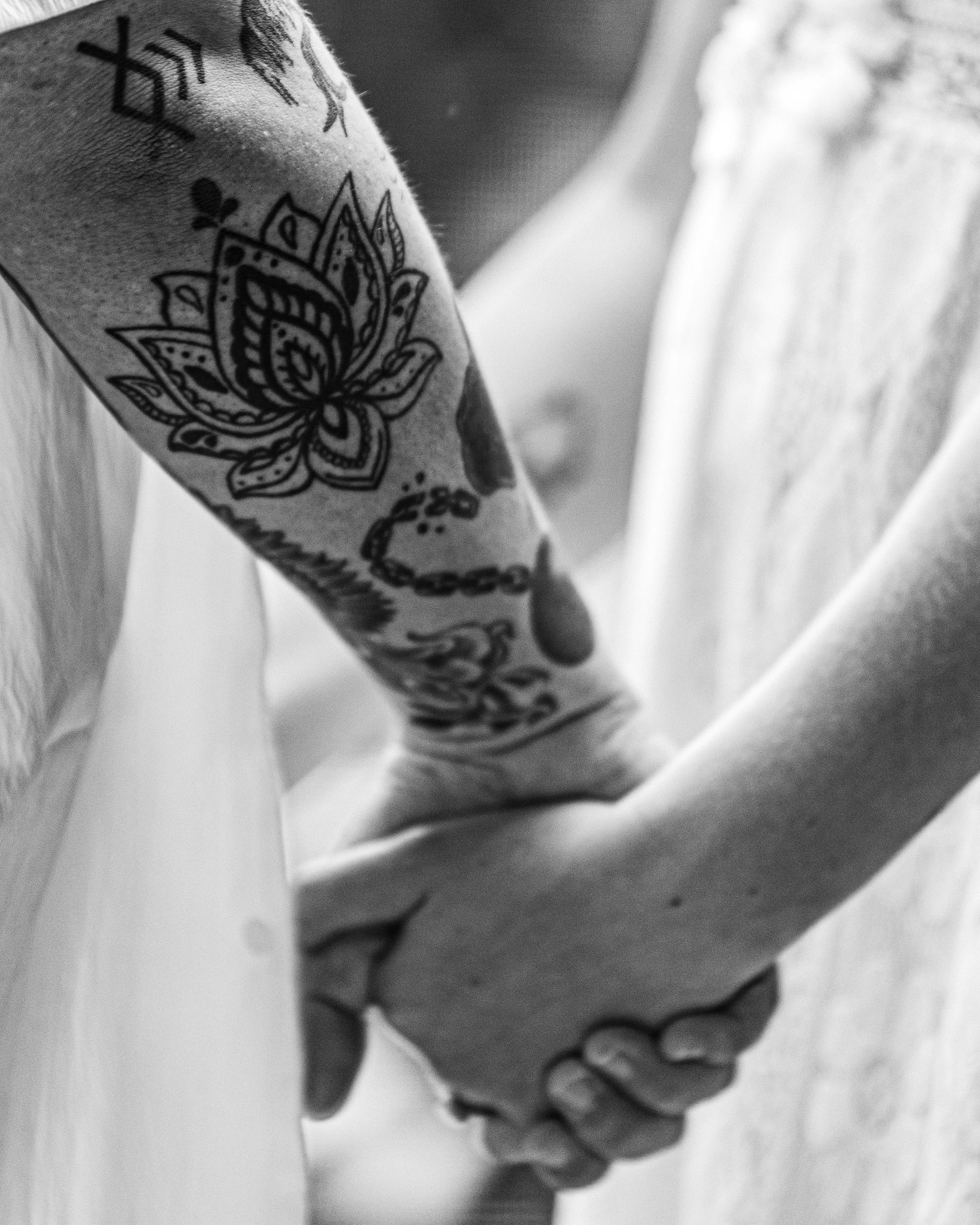 A close-up black-and-white photo of two people holding hands, one with tattoos on their arm.