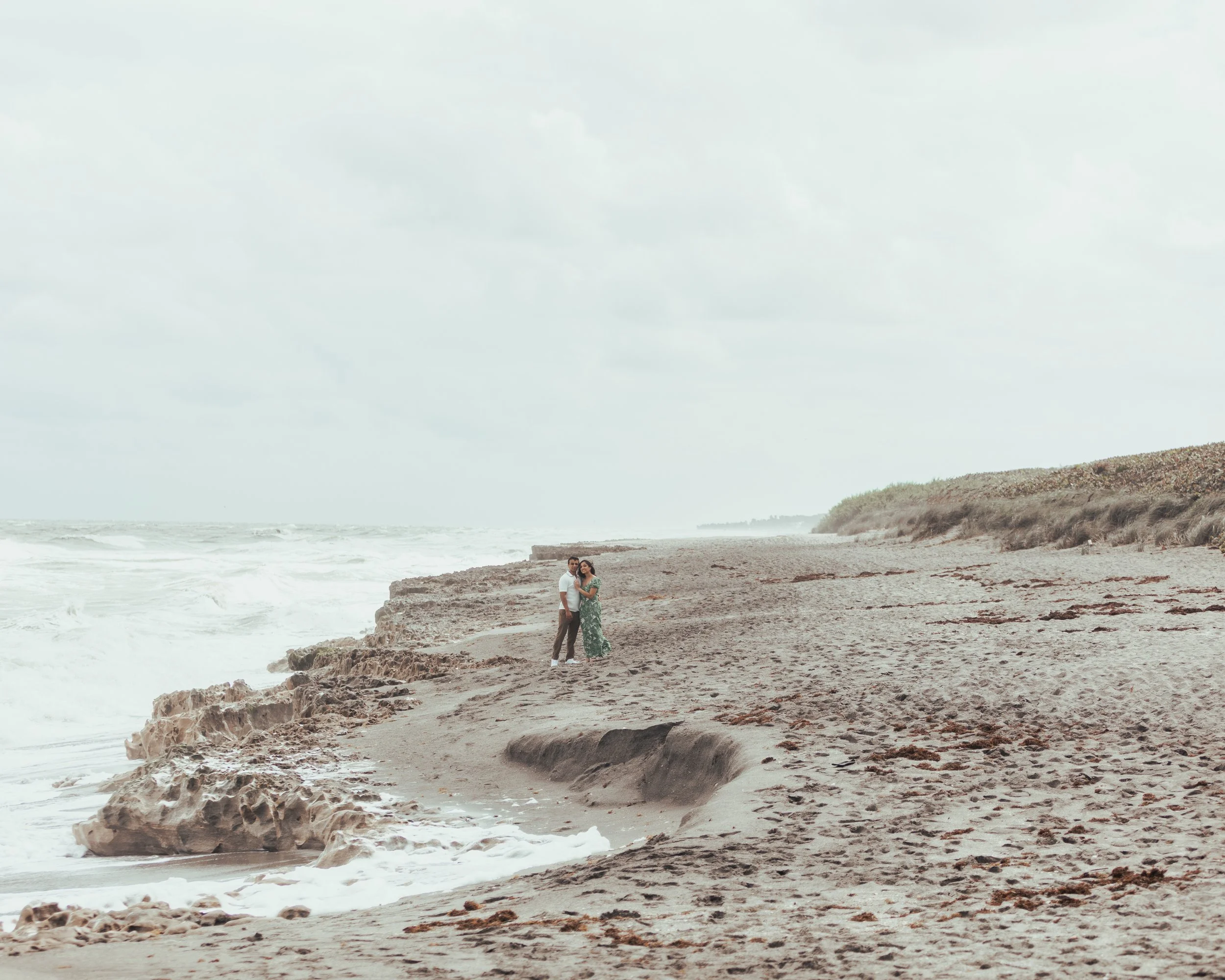 A couple standing on a rocky beach near the ocean with sand dunes in the background.