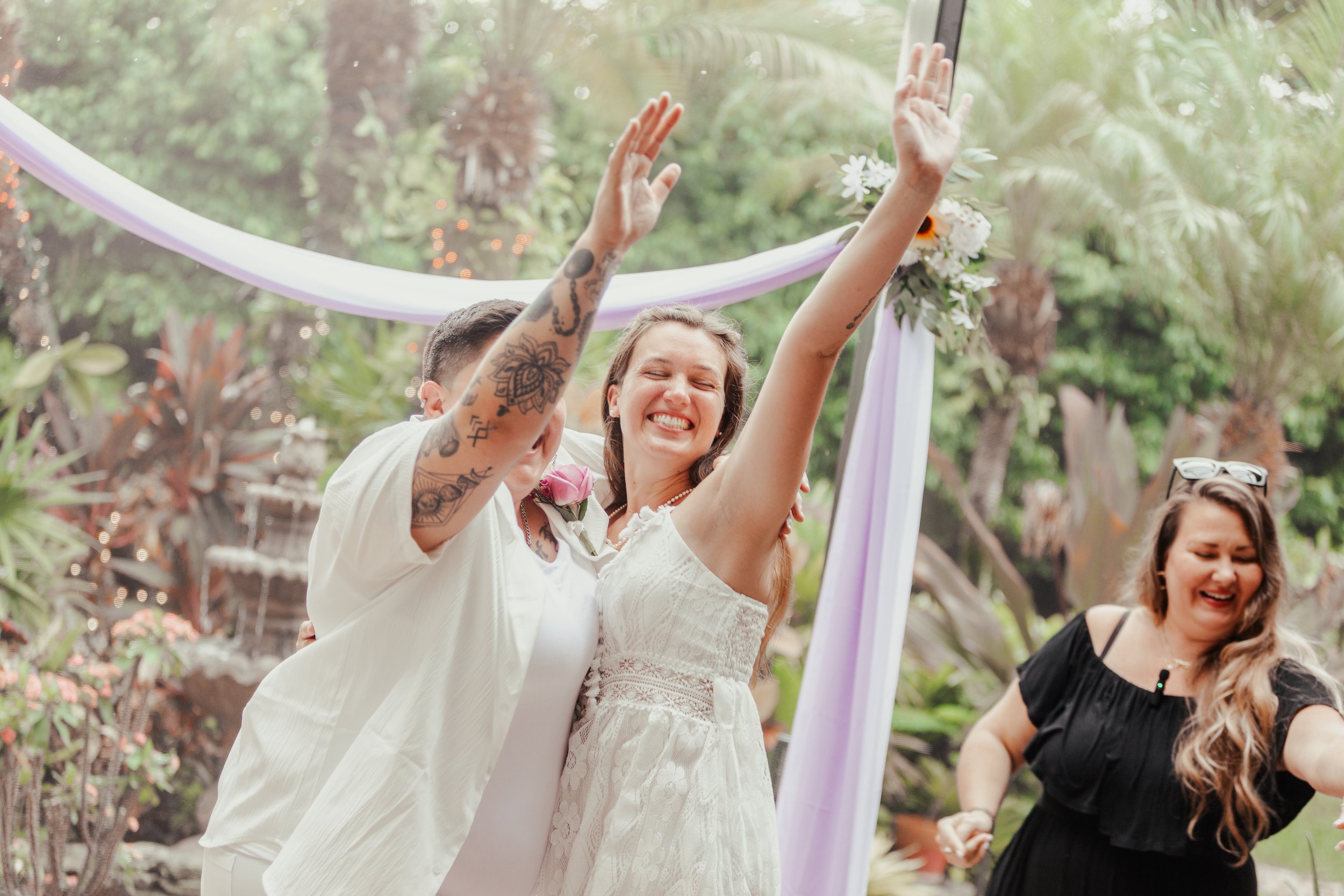 People celebrating wedding outdoors, smiling, raising hands, purple floral arch, greenery background