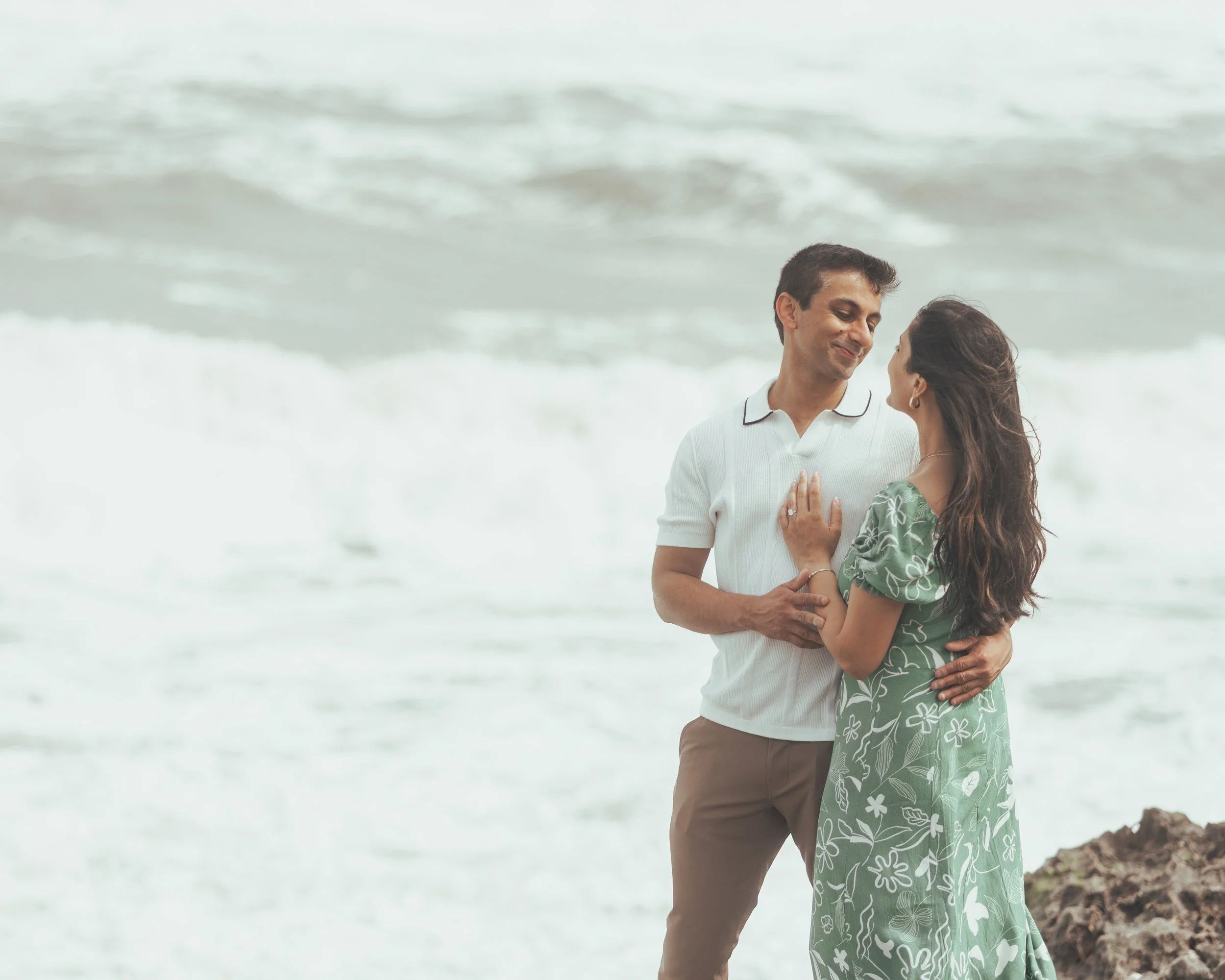 A couple standing on a rocky beach, facing each other and sharing a tender moment as waves crash behind them.