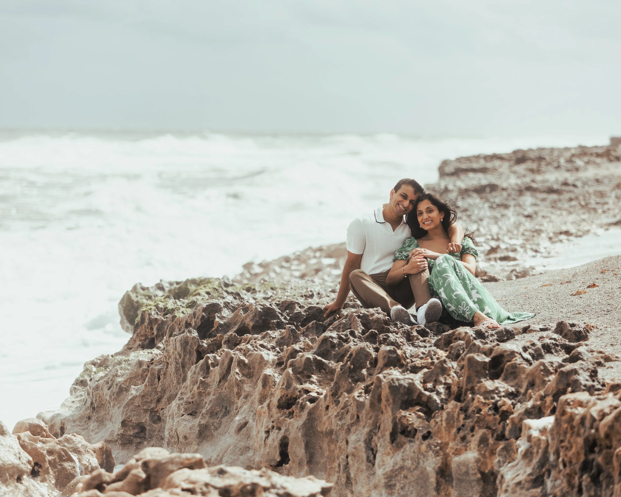 A happy couple sitting on rocks on a beach, embracing and smiling, with the ocean and waves in the background.