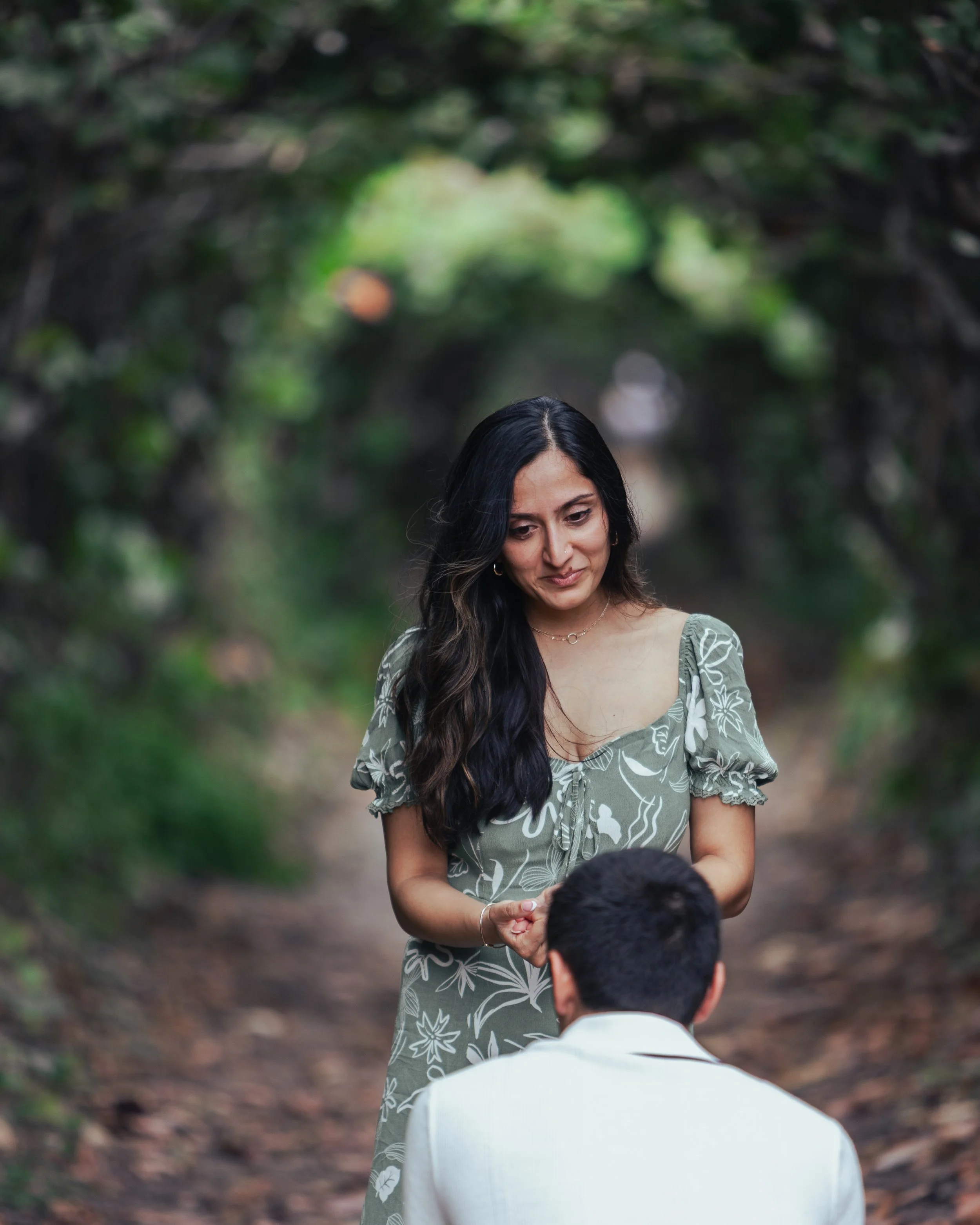 A woman with long dark hair in a floral dress kneeling in front of a man with short dark hair in a white suit, outside on a wooded path.