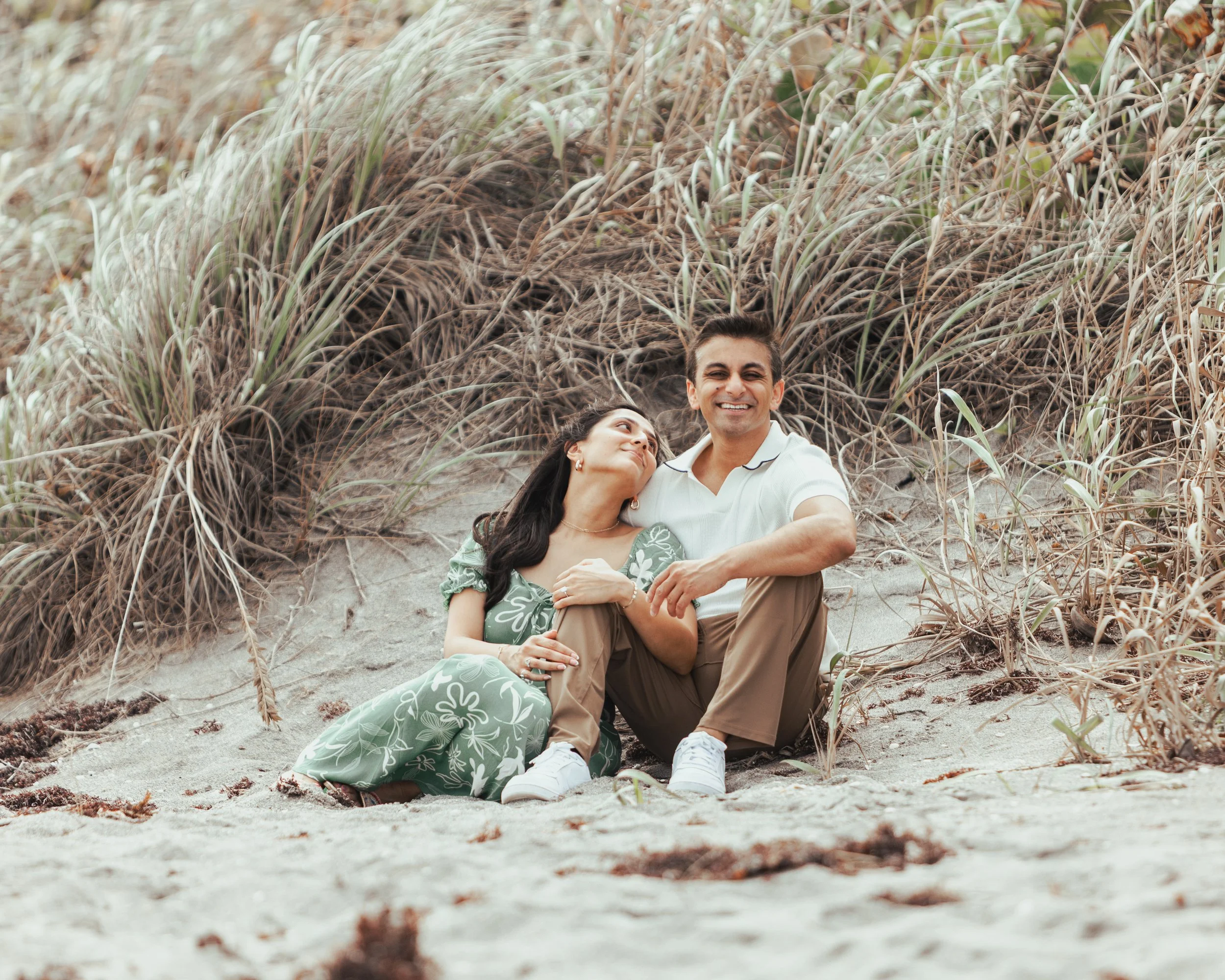 A happy couple sitting on a sandy beach with tall grasses and bushes behind them, smiling and enjoying each other's company.