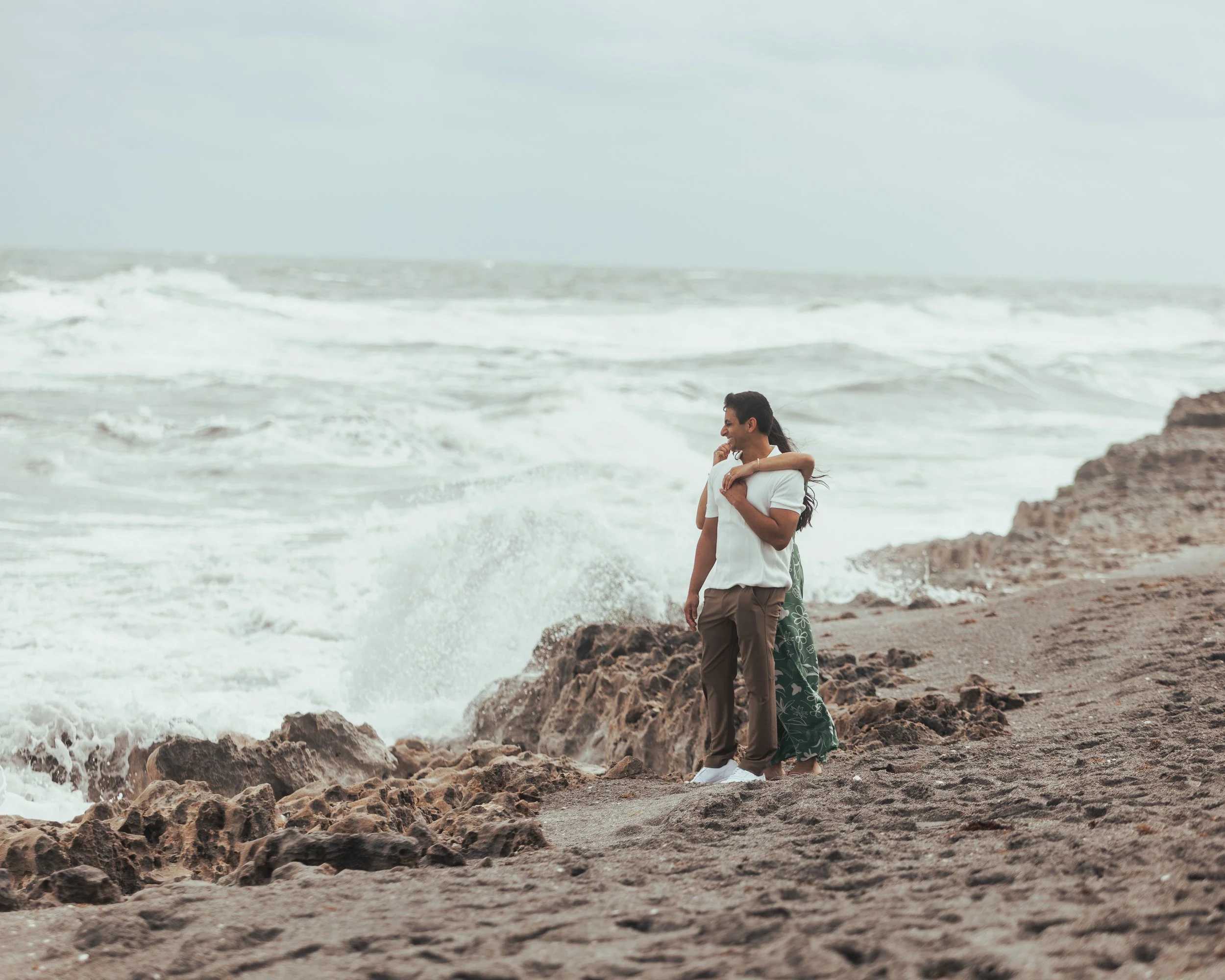 A couple hugging on a rocky beach with waves crashing behind them.