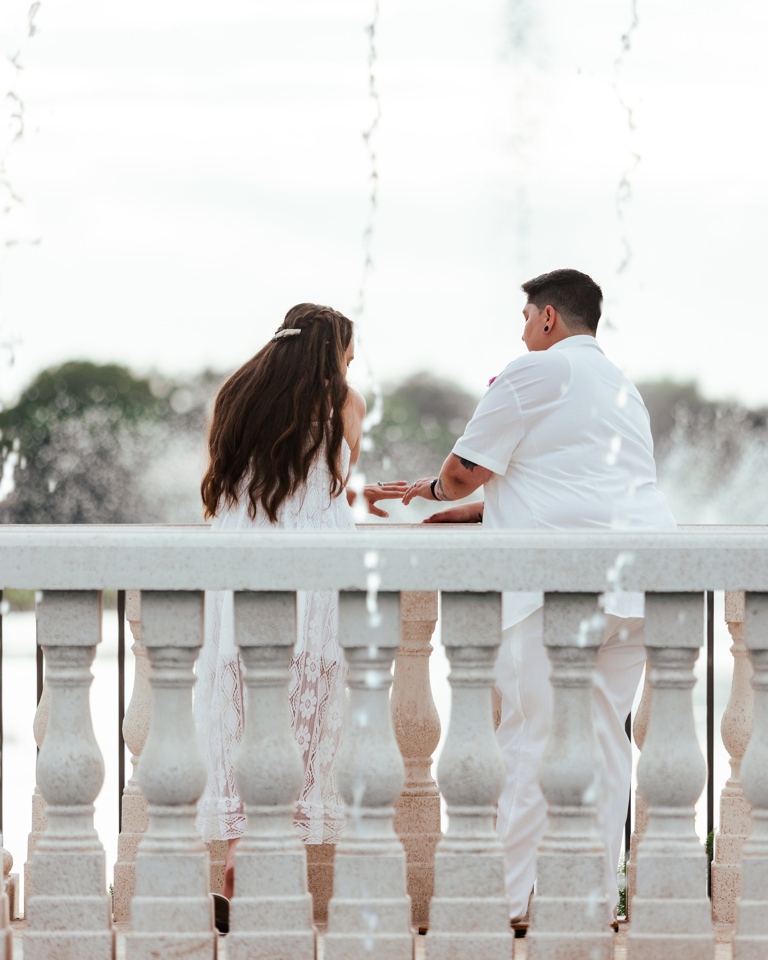 A couple sitting on a balcony with water fountains, sharing a moment during a wedding or special occasion, with cloudy sky and trees in the background.