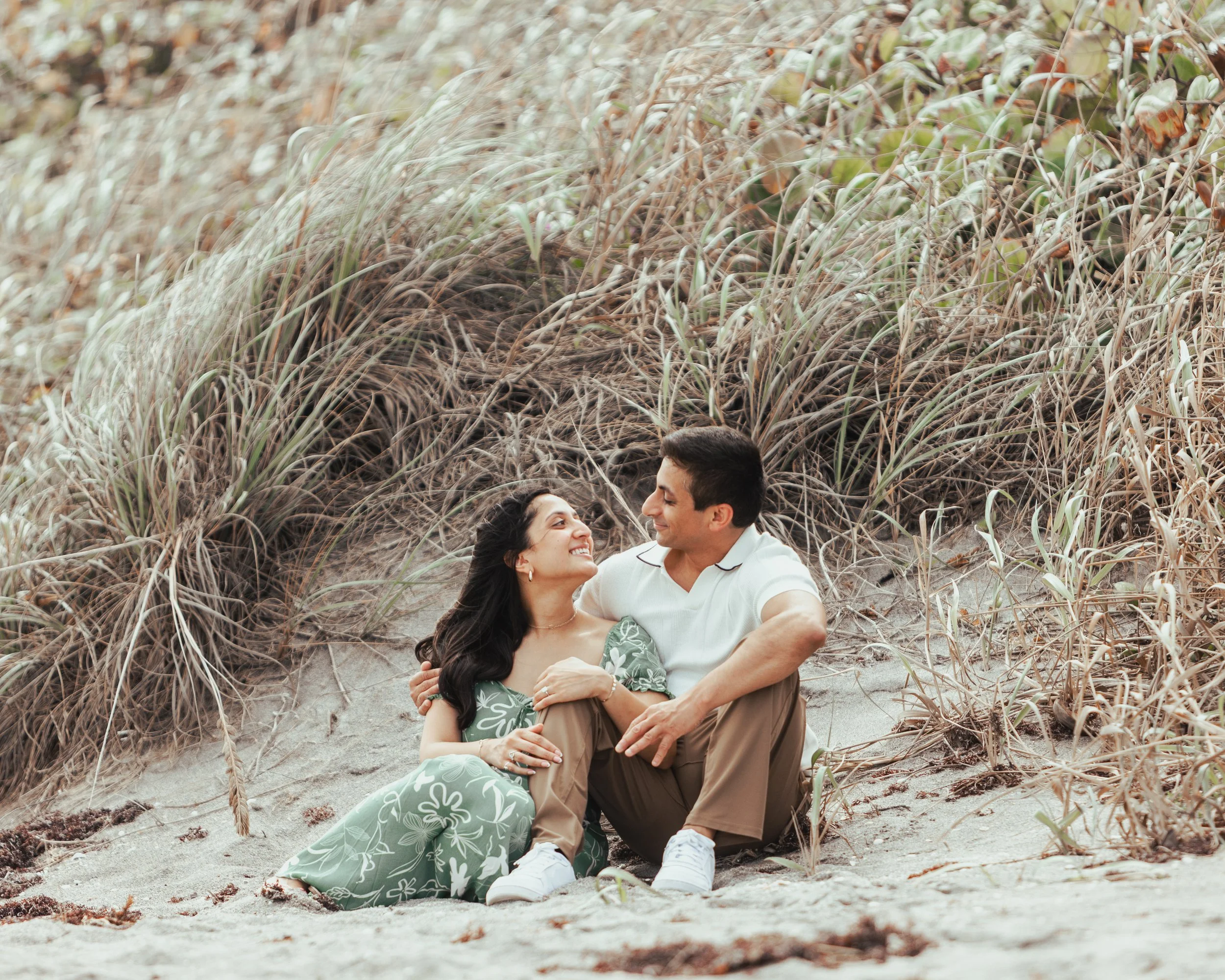 A couple sitting on sandy beach with tall grass behind them, smiling and looking at each other.