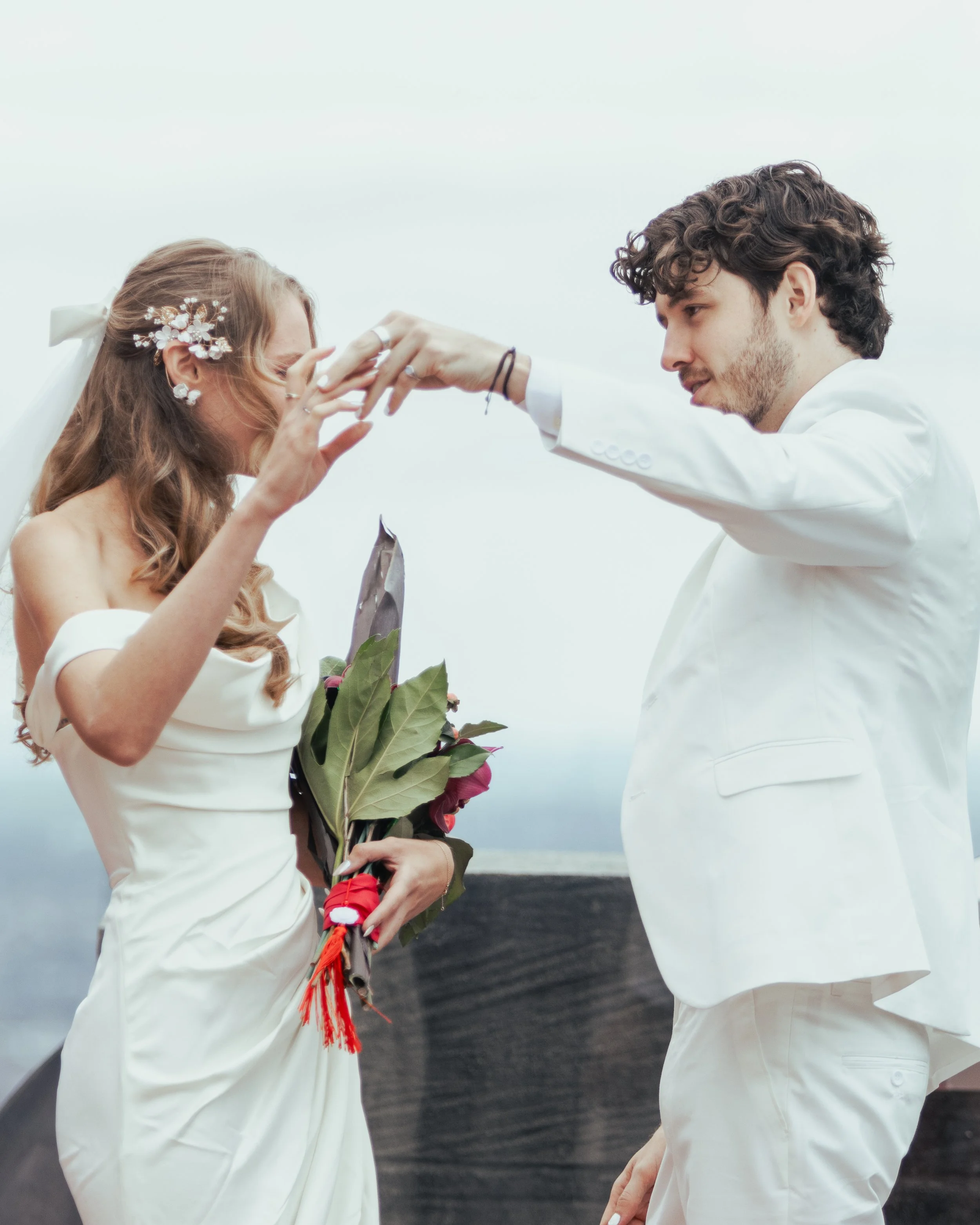 Couple exchanging wedding rings on a cloudy day, with the bride holding a bouquet of flowers.