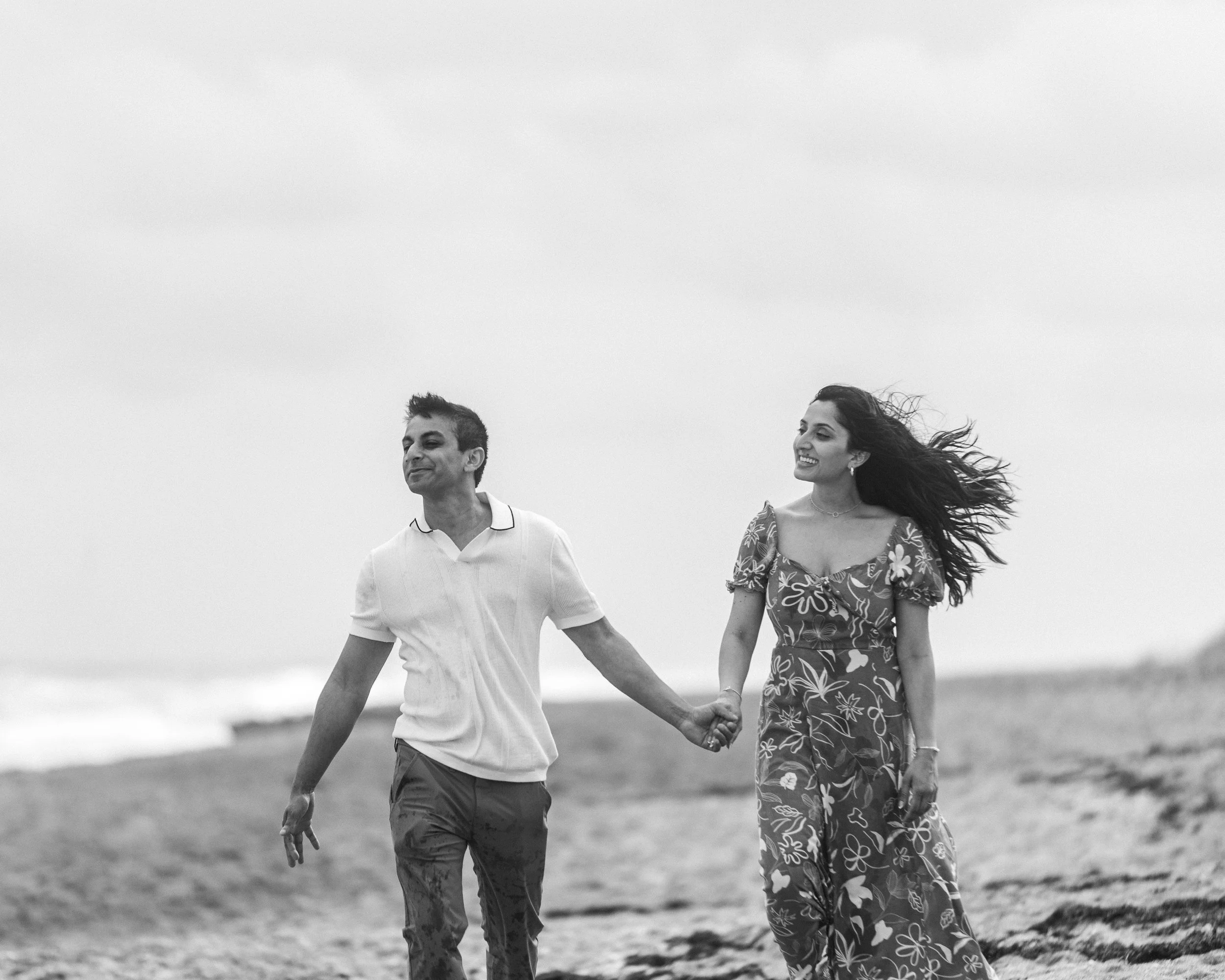 A couple walking hand-in-hand on the beach, smiling, with the woman’s hair blowing in the wind.