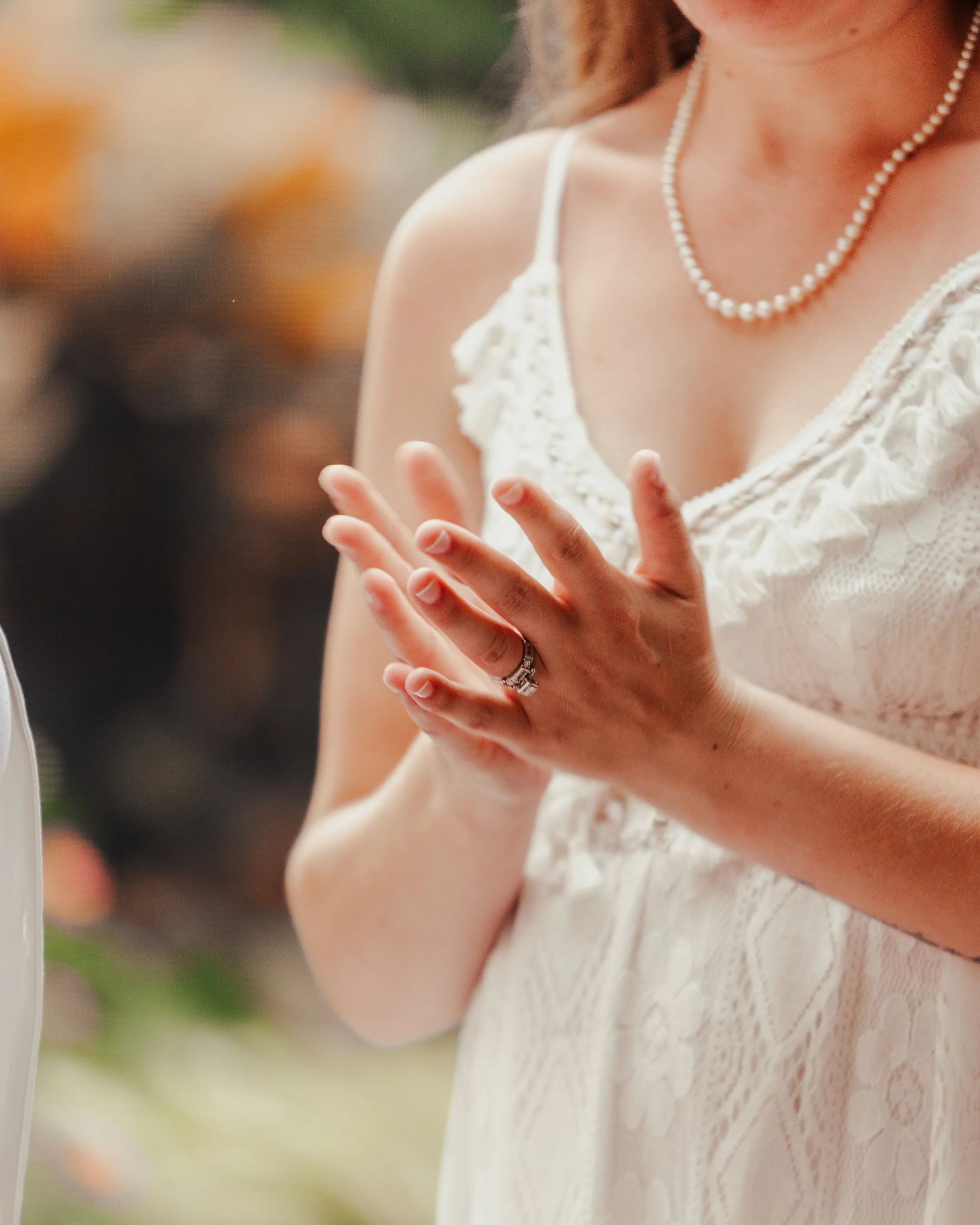 A woman wearing a white lace dress and a pearl necklace, with her hands clasped together near her chest, displaying a wedding ring.