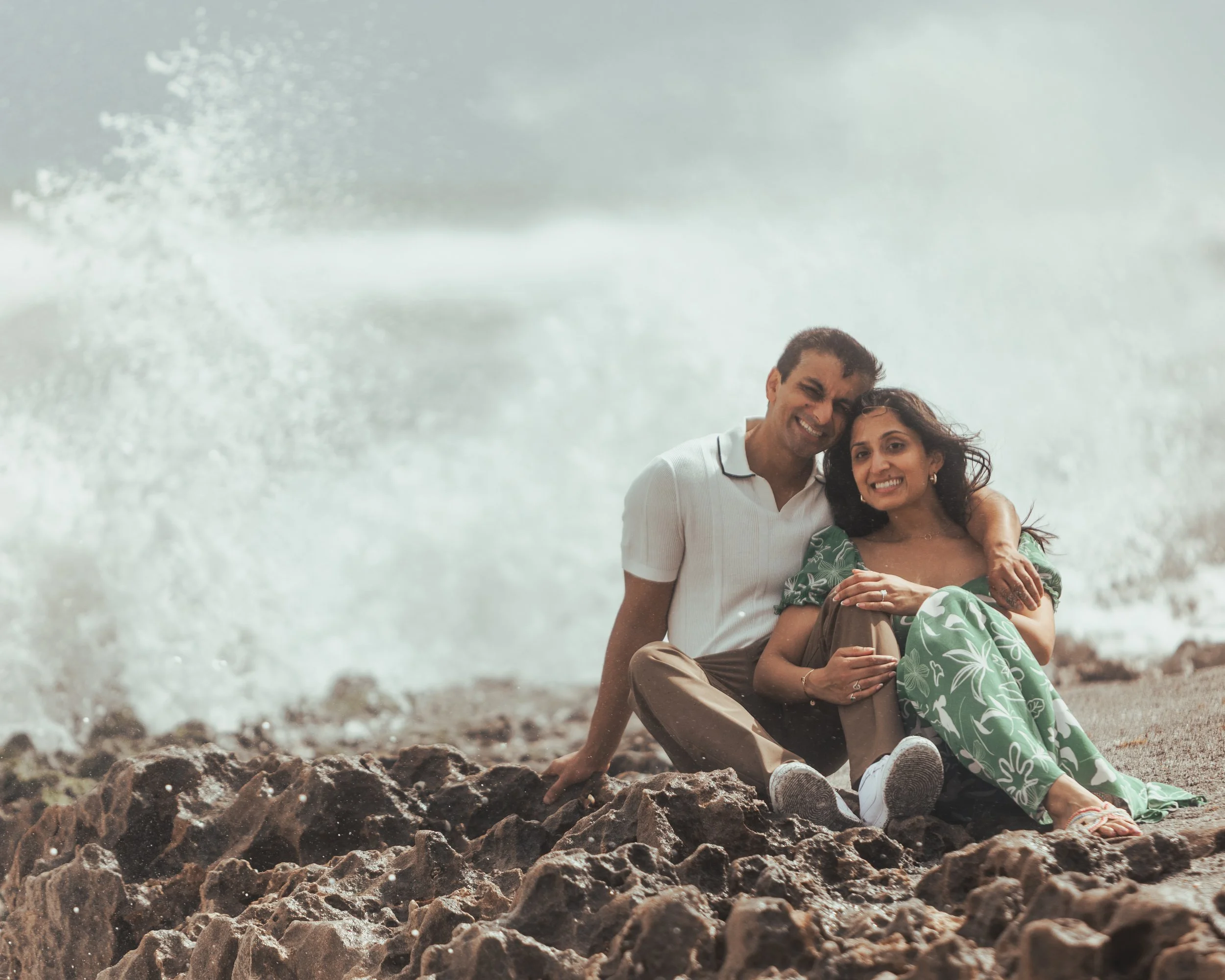 A smiling couple sitting on rocks near the ocean, with waves crashing behind them.