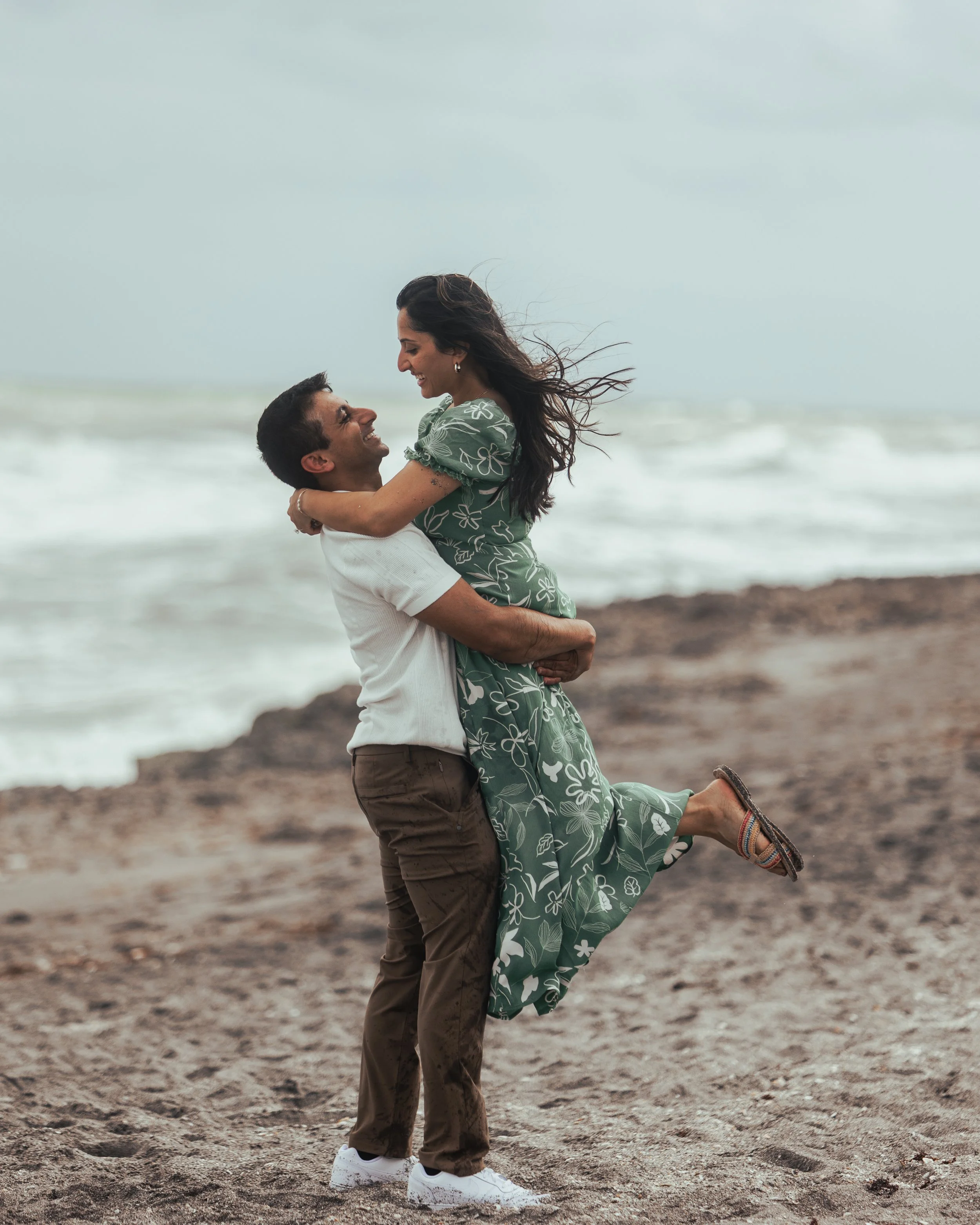 A happy couple on the beach with the man lifting the woman in his arms, both smiling and looking at each other, with ocean waves and cloudy sky in the background.