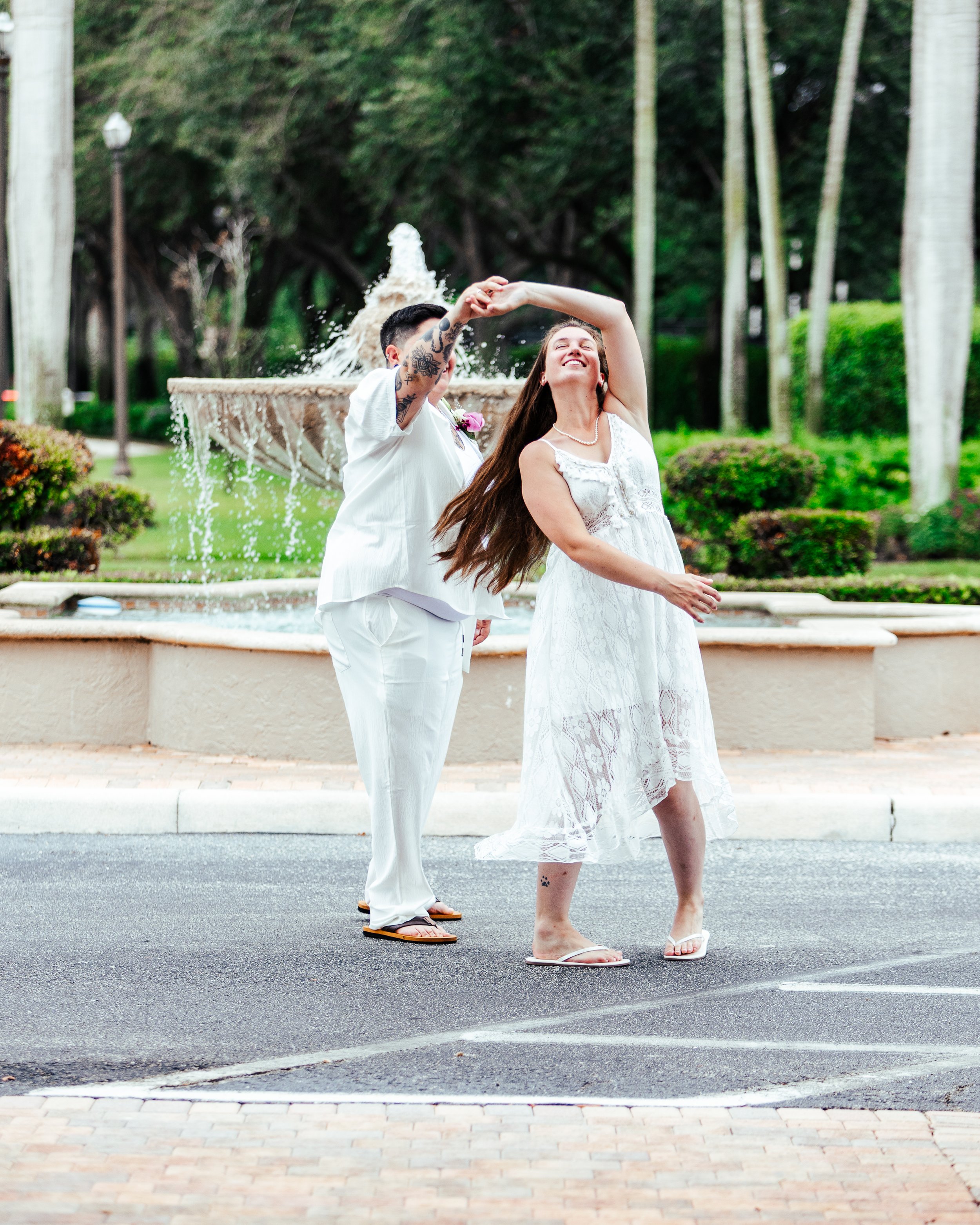 A couple dancing in front of a fountain in a park, with trees and bushes in the background.