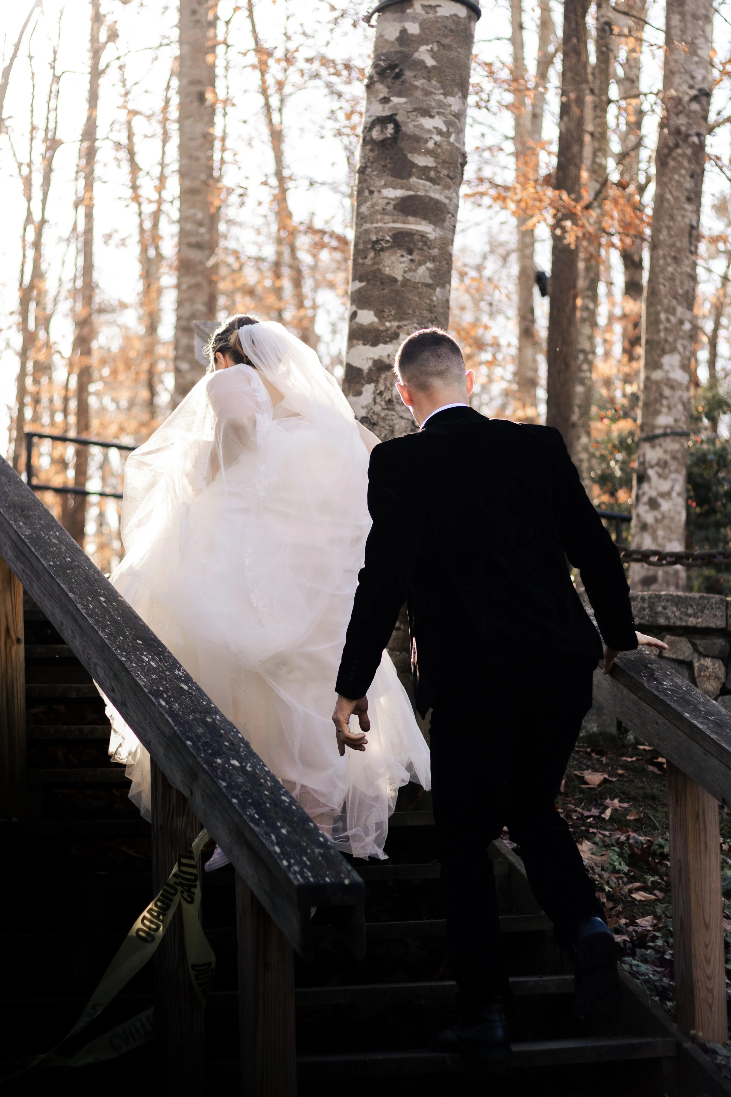 A bride and groom ascending outdoor wooden stairs in a wooded area during daylight, with the bride in a white wedding dress and veil, and the groom in a black suit.