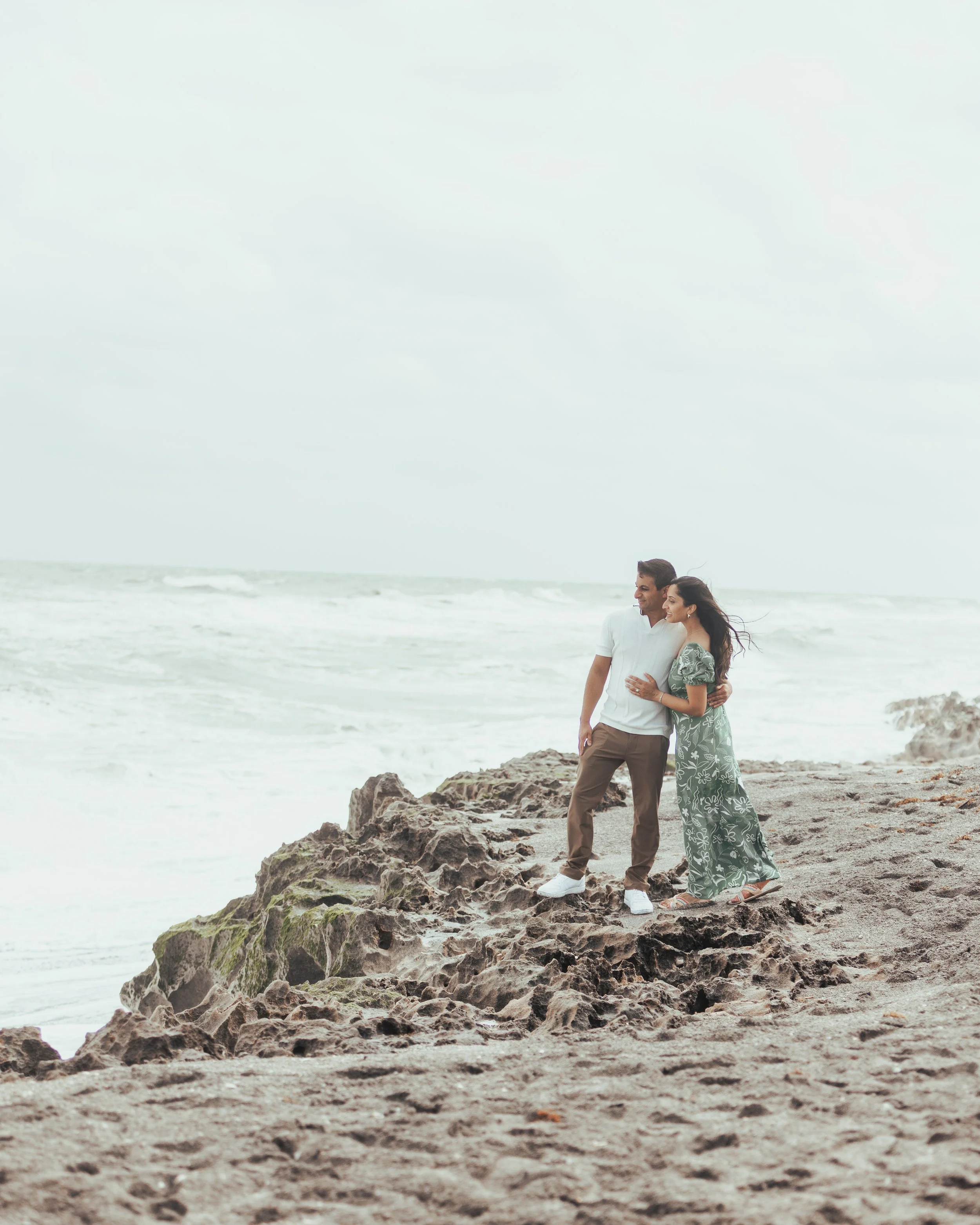 Couple standing on rocky beach near the ocean, wind in hair, casual clothing, cloudy sky.