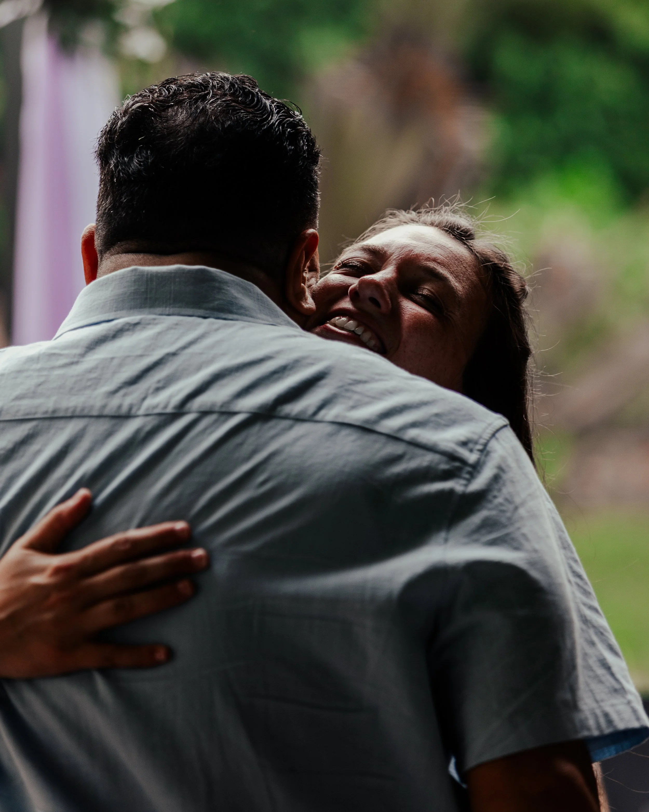 A woman and a man embrace, smiling, outdoors with blurred greenery in the background.