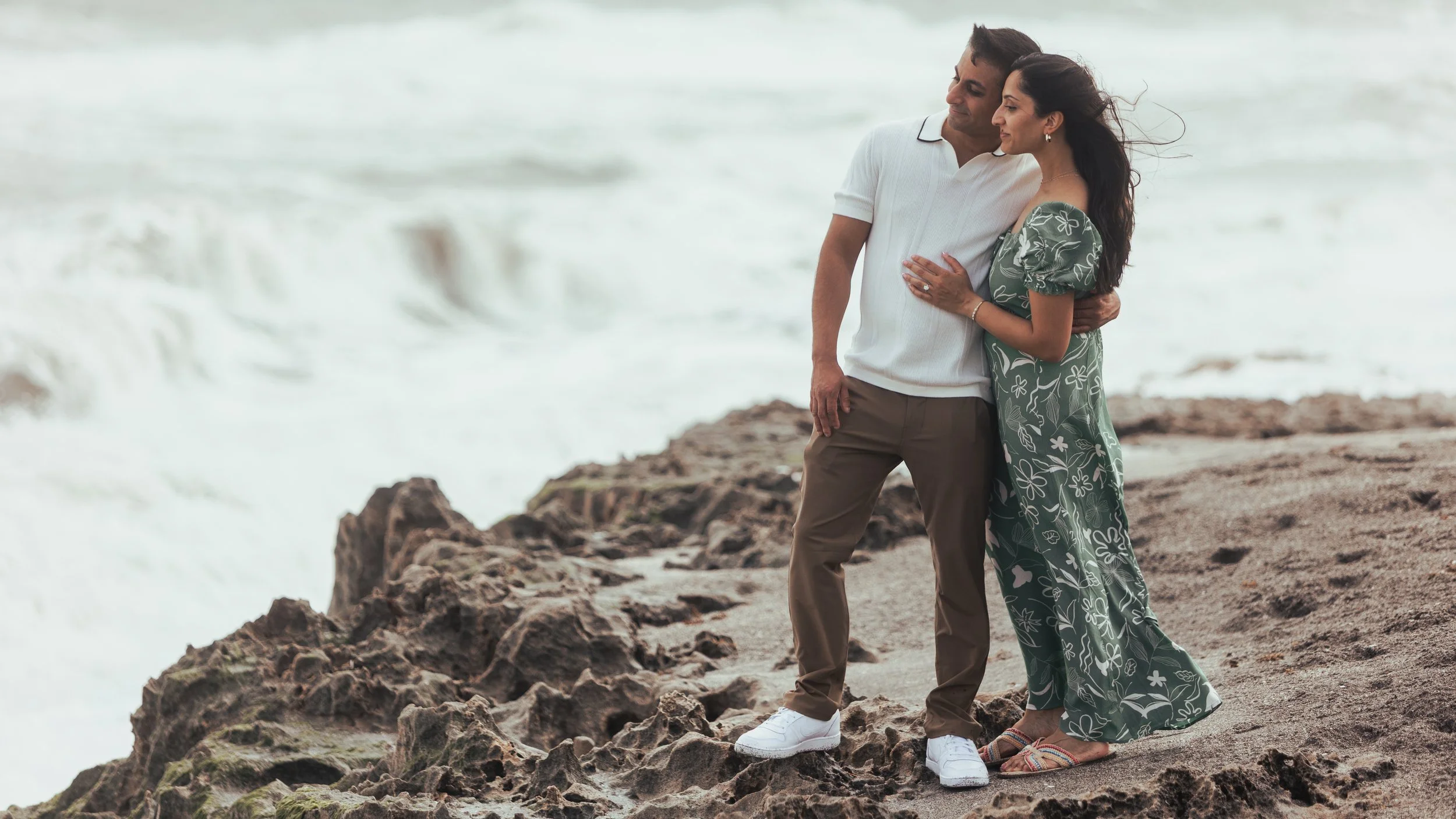 A couple standing close together on a rocky beach with ocean waves in the background. The man is in a white shirt and khaki pants, and the woman is in a green floral dress, both looking at each other.
