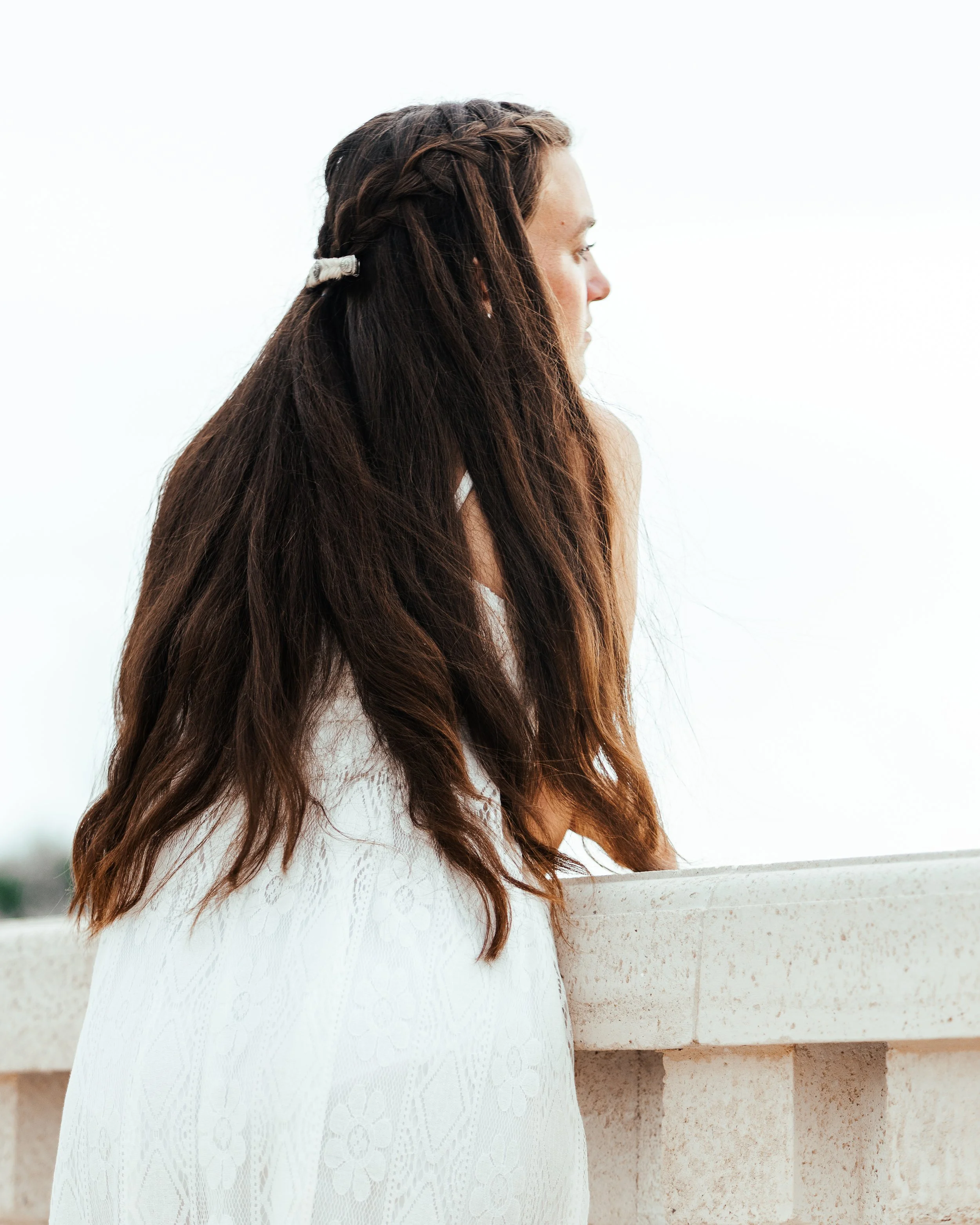 A woman with long, wavy brown hair and a side braid, wearing a white dress, standing next to a beige stone wall and gazing to the right.